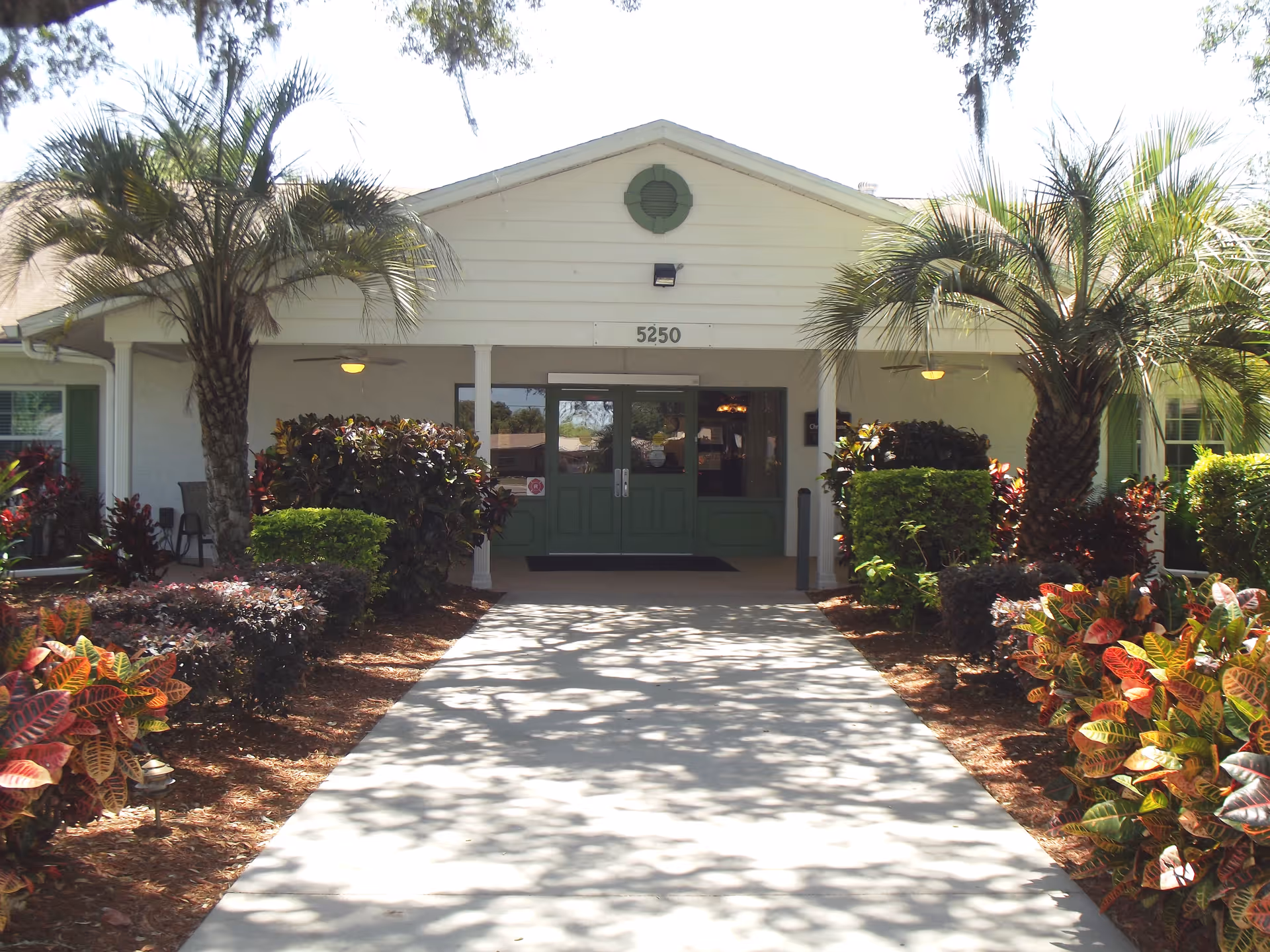 Front entrance of a single-story building with a paved walkway, green double doors, palms and landscaped shrubs.