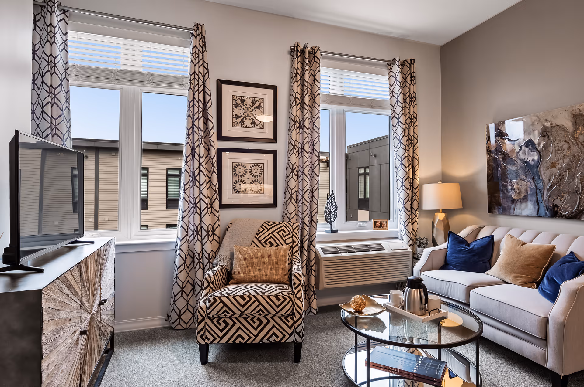 A cozy living room with a patterned armchair and a beige sofa adorned with blue and tan pillows. There is a glass coffee table with a tray holding a coffee pot and cups. The room has two large windows with geometric patterned curtains, a TV on a wooden cabinet, a table lamp, and abstract wall art.