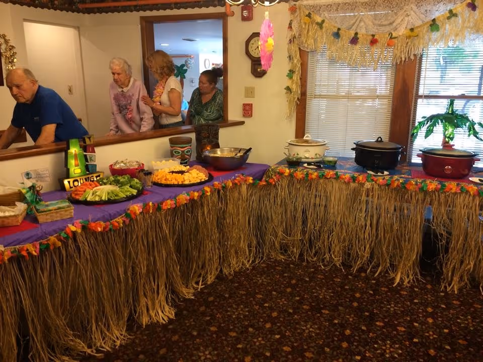 A buffet table decorated with a tropical theme, including grass skirts and colorful flower garlands. The table is covered with a purple cloth and holds trays of vegetables, cheese cubes, and several slow cookers. In the background, four people are seen near a pass-through window, with one man leaning on the counter and three women standing behind him. The room has windows with blinds and a potted plant on the windowsill.