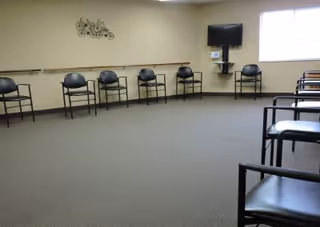 Empty activity room with chairs arranged around the perimeter, a wall-mounted TV, and a large window.