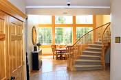 Interior view of a senior living facility with a wooden staircase on the right, a round dining table with chairs near large windows letting in natural light, and a wooden door on the left.
