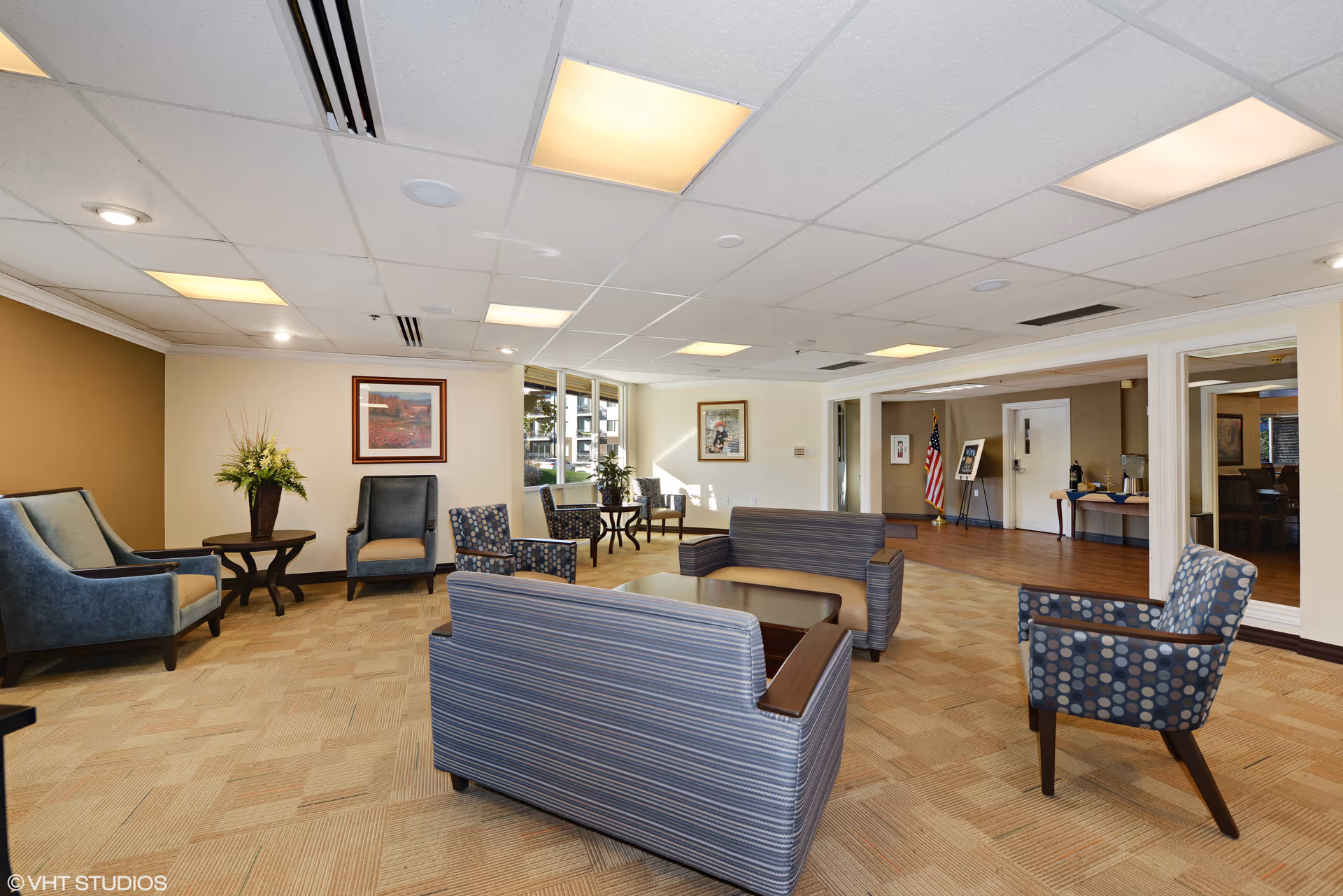 A spacious seating area with upholstered chairs and sofas arranged around tables in a bright senior living facility lounge.