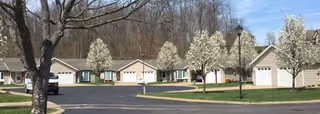 Row of single-story cottage-style senior living units with attached garages, blossoming trees, and a paved driveway.