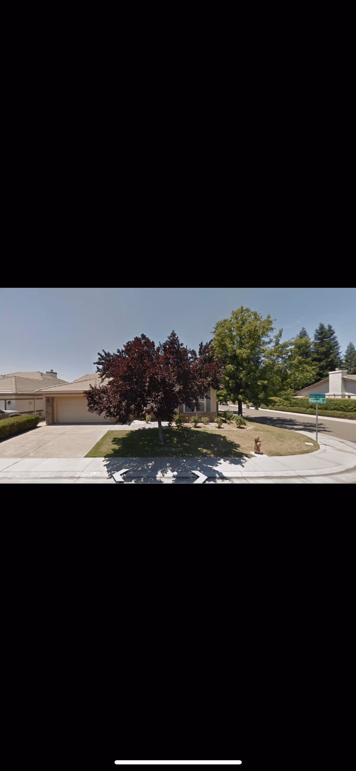 A suburban residential street corner with a single-story house featuring a two-car garage. There are two trees in the front yard, one with dark red leaves and the other with green leaves. A fire hydrant is visible near the curb, and a street sign is present at the corner. The sky is clear and blue.