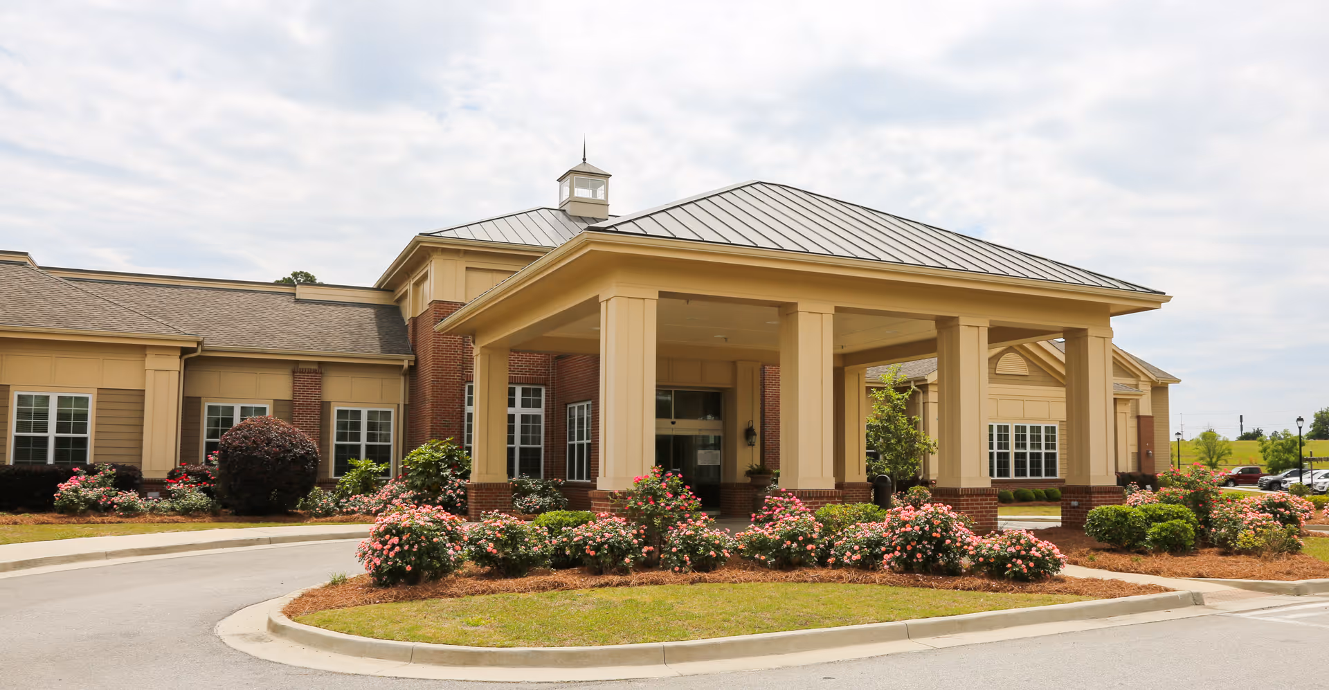 Exterior view of Meadows Park Health & Rehabilitation building entrance with a covered drop-off area, surrounded by landscaped bushes and flowers under a partly cloudy sky.