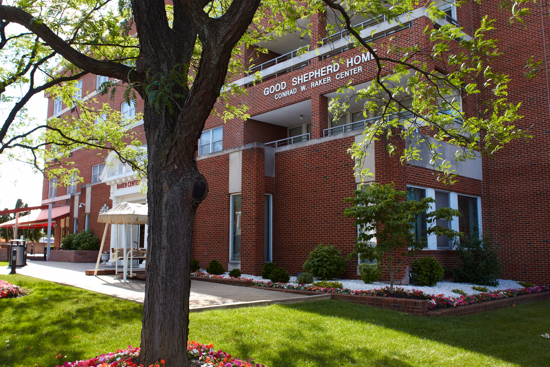 Exterior view of Good Shepherd Home - Conrad W. Raker Center, a multi-story red brick building with balconies. In the foreground, there is a large tree with green leaves and a well-maintained lawn with flower beds surrounding the building.