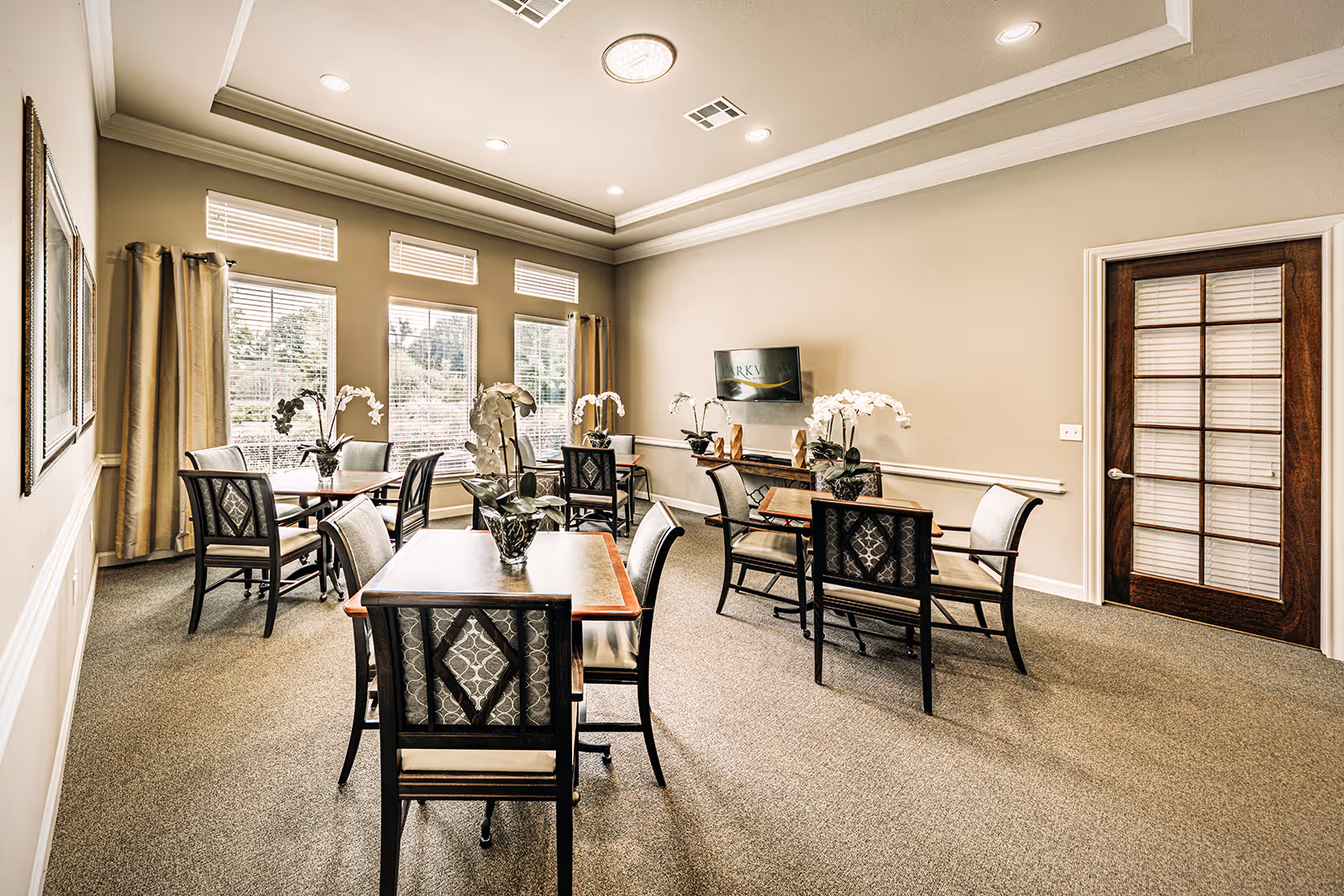 A bright and spacious dining room with several wooden tables and cushioned chairs arranged neatly. Each table has a decorative white orchid centerpiece. Large windows with beige curtains allow natural light to fill the room. A flat-screen TV is mounted on the far wall above a wooden console. The room has beige walls, a carpeted floor, and a wooden door with glass panels on the right side.