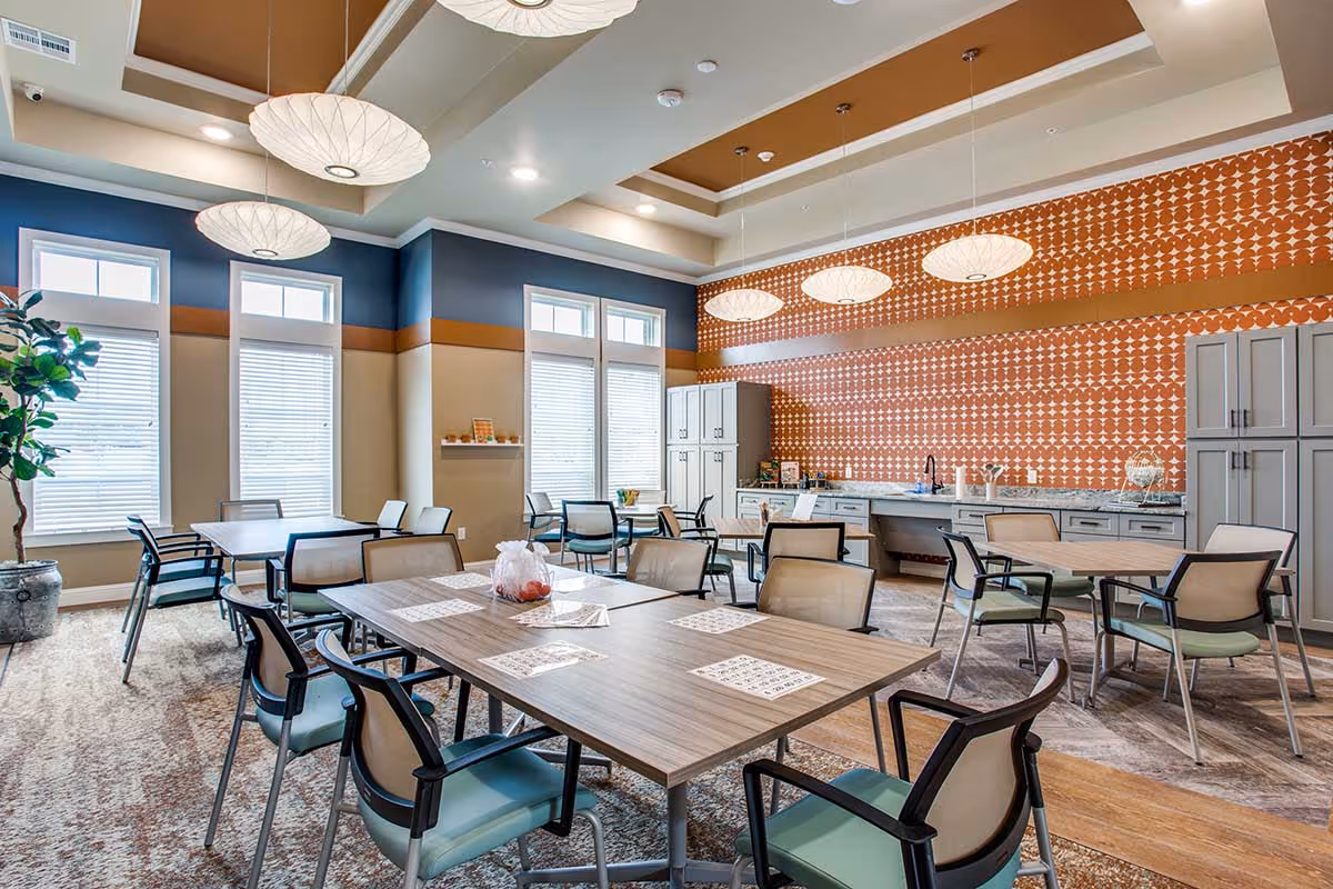 A bright and spacious dining area with multiple tables and chairs arranged neatly. The room features large windows with blinds, a patterned orange accent wall with cabinets and a countertop, and modern hanging light fixtures. There is a potted plant in the corner and a mix of carpet and wood flooring.