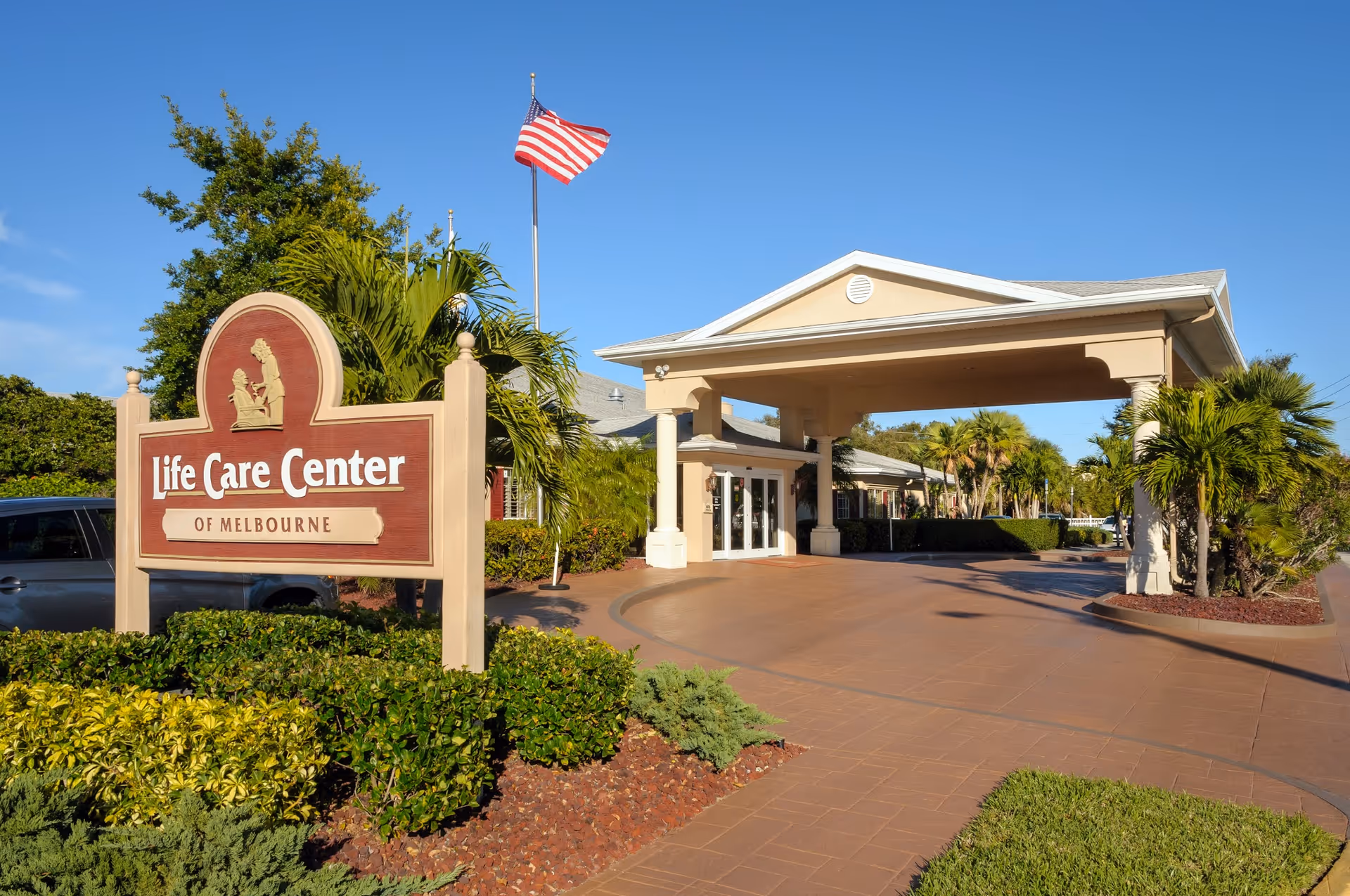 Front entrance of Life Care Center of Melbourne with a wooden sign, covered porte-cochere, American flag and landscaped grounds.