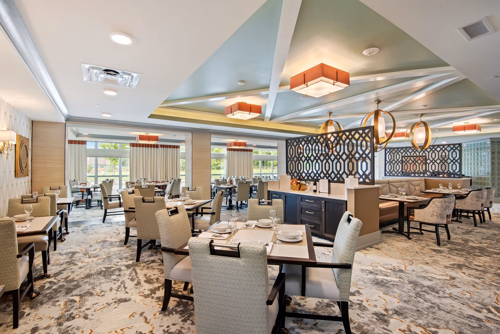 A spacious and well-lit dining room in a senior living facility with multiple tables and chairs arranged neatly. The room features modern light fixtures, decorative partitions, large windows with curtains allowing natural light, and a patterned carpeted floor. Tables are set with plates, glasses, and napkins, ready for dining.