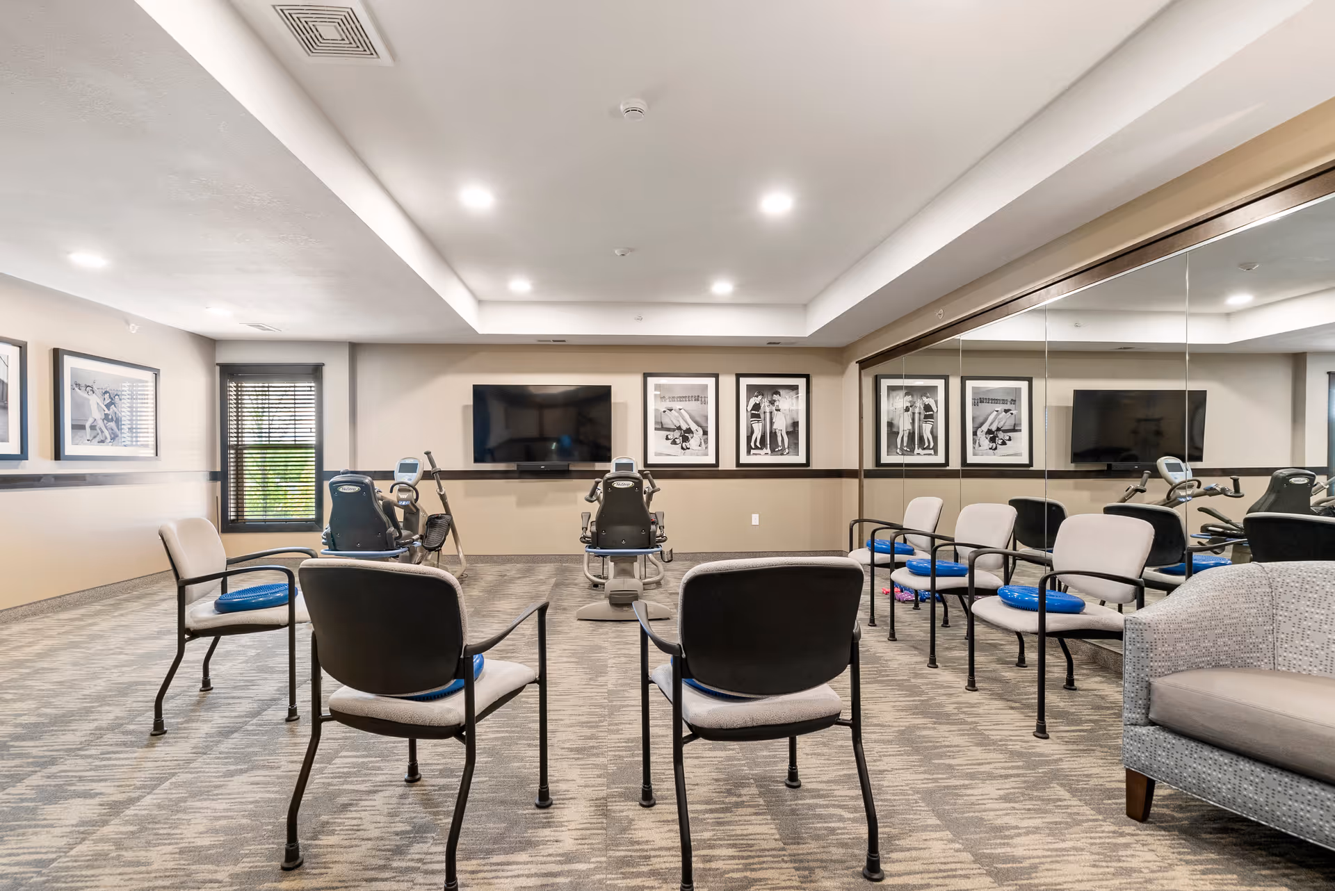 A senior living facility exercise room with chairs arranged in a circle, two recumbent exercise bikes facing a wall-mounted TV, framed black and white photos on the walls, a large mirror covering one wall, and a window with blinds letting in natural light.