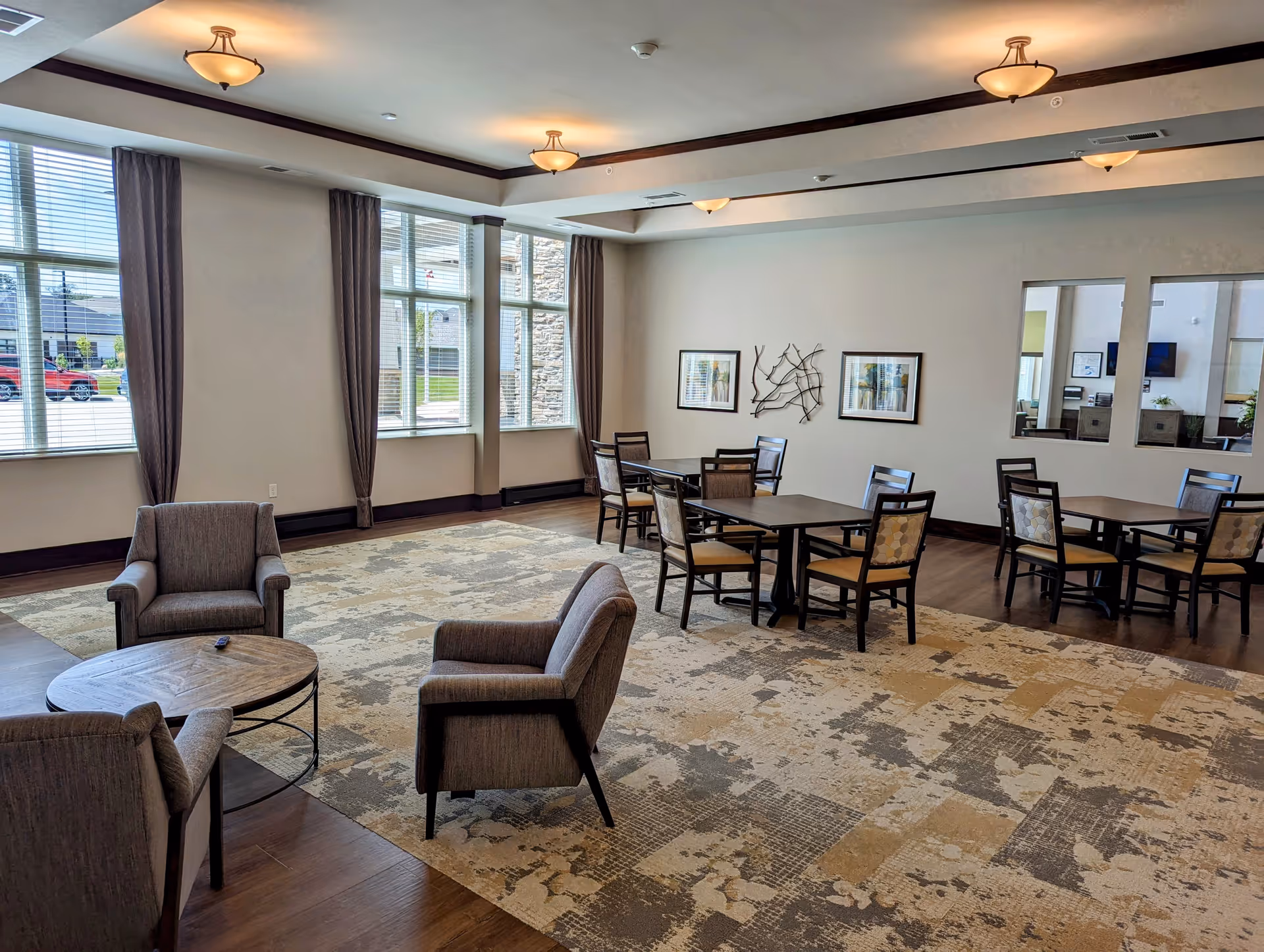 A spacious common area in a senior living facility with large windows letting in natural light. The room features a patterned carpet, three gray upholstered armchairs around a round wooden coffee table, and several square tables with chairs arranged for dining or socializing. The walls are decorated with framed artwork and a metal wall sculpture. Ceiling lights provide additional illumination.