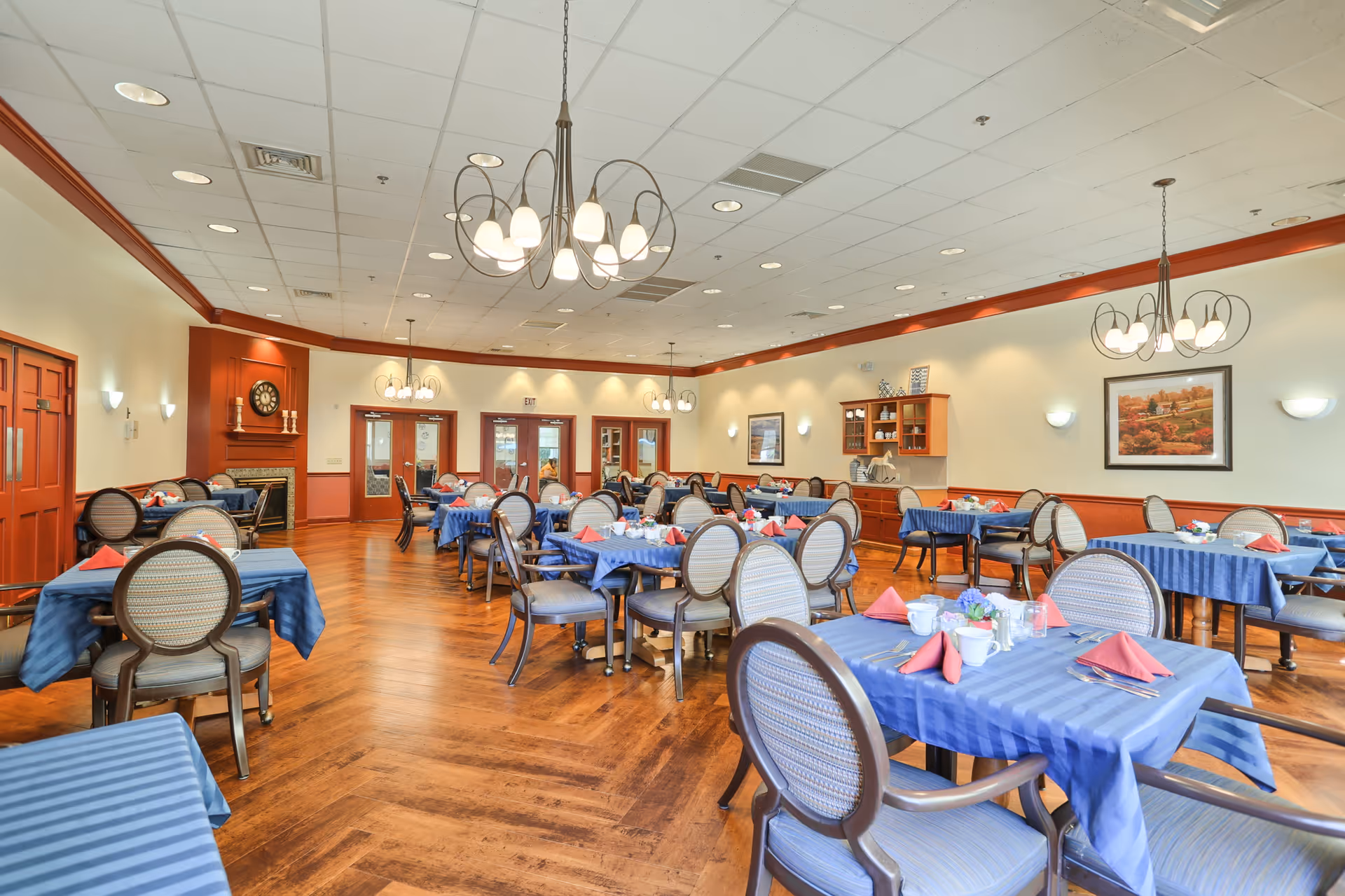 A spacious dining room with multiple tables covered in blue tablecloths and set with red napkins, white cups, and silverware. The room features wooden floors, beige walls with red trim, several chandeliers hanging from a white tiled ceiling, and framed artwork on the walls. There are cushioned chairs around each table and a cabinet with decorative items on the right side of the room.