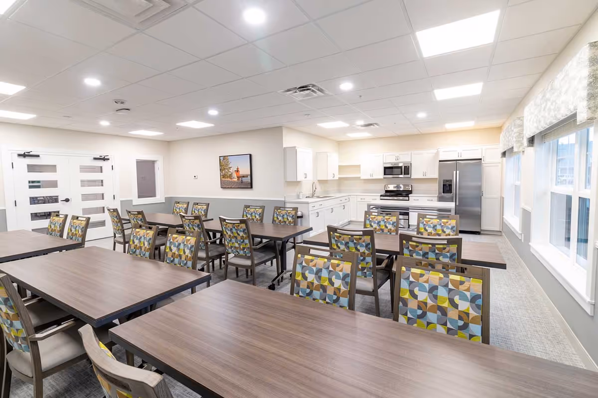 A bright and spacious dining area with multiple wooden tables and chairs featuring colorful geometric patterned upholstery. The room has large windows with valances, white walls, and a ceiling with recessed lighting. In the background, there is a modern kitchen area with white cabinets, stainless steel appliances including a refrigerator, oven, and microwave, and a sink.