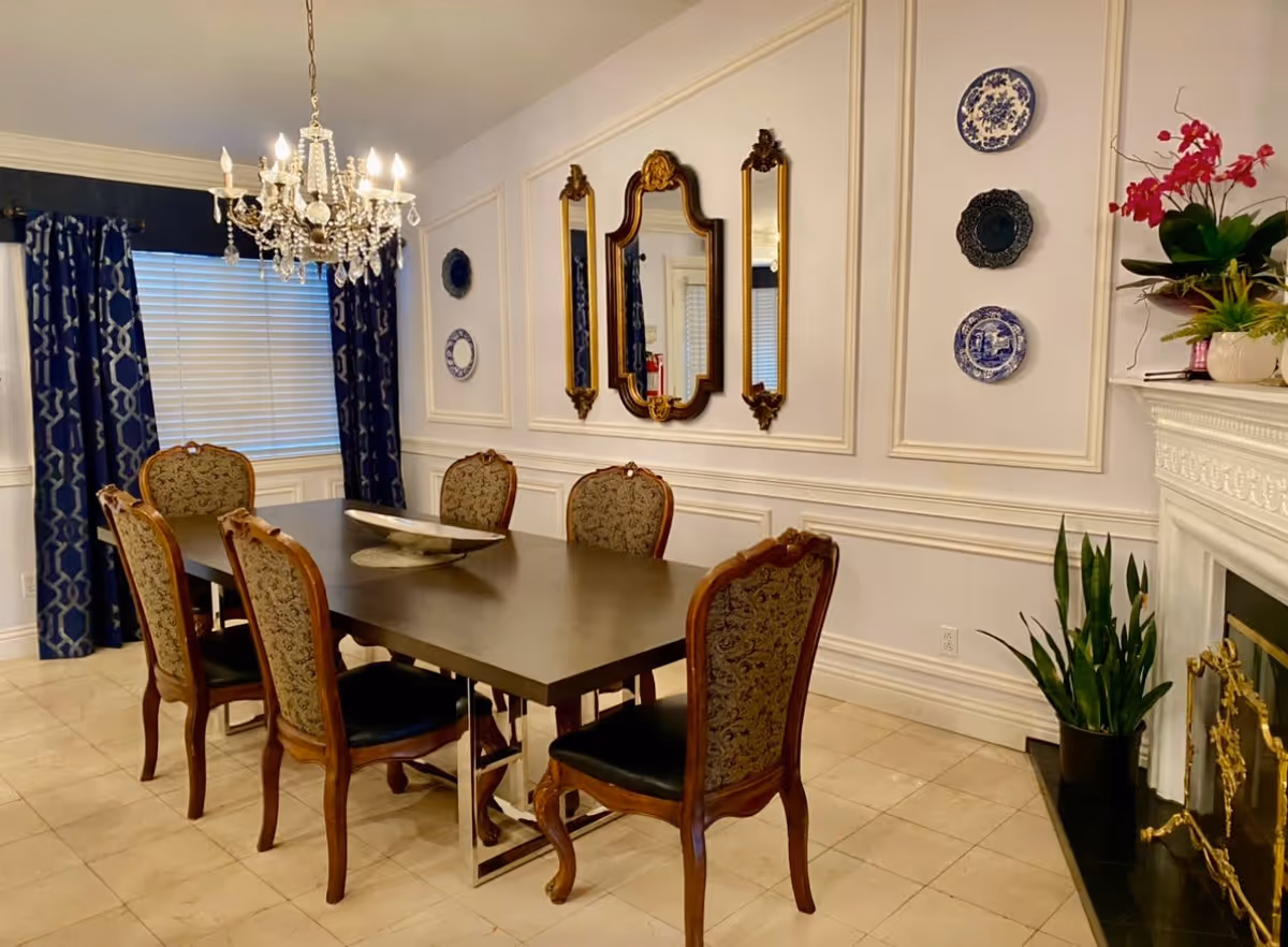 Elegant dining room with a rectangular dark wood table surrounded by six upholstered chairs with ornate wooden frames. A crystal chandelier hangs above the table. The walls are decorated with three mirrors and three decorative plates. There are dark blue patterned curtains on the window and a fireplace with plants and flowers on the mantel.