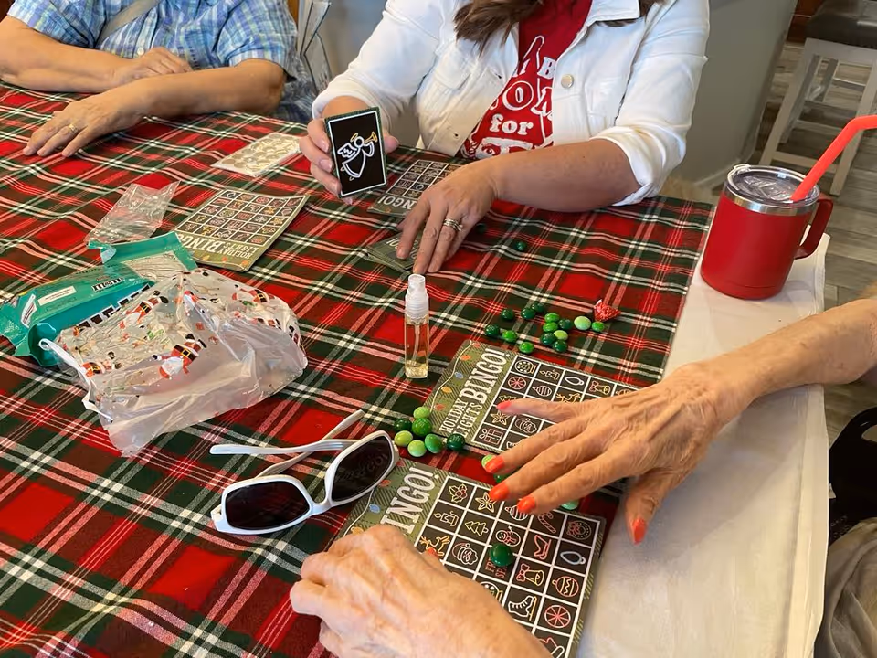 Three people sitting around a table covered with a red and green plaid tablecloth playing holiday-themed bingo. One person is holding a bingo card with an angel illustration, while the others have bingo cards with green candies placed on them. There are also sunglasses, a small spray bottle, and a red cup with a straw on the table.