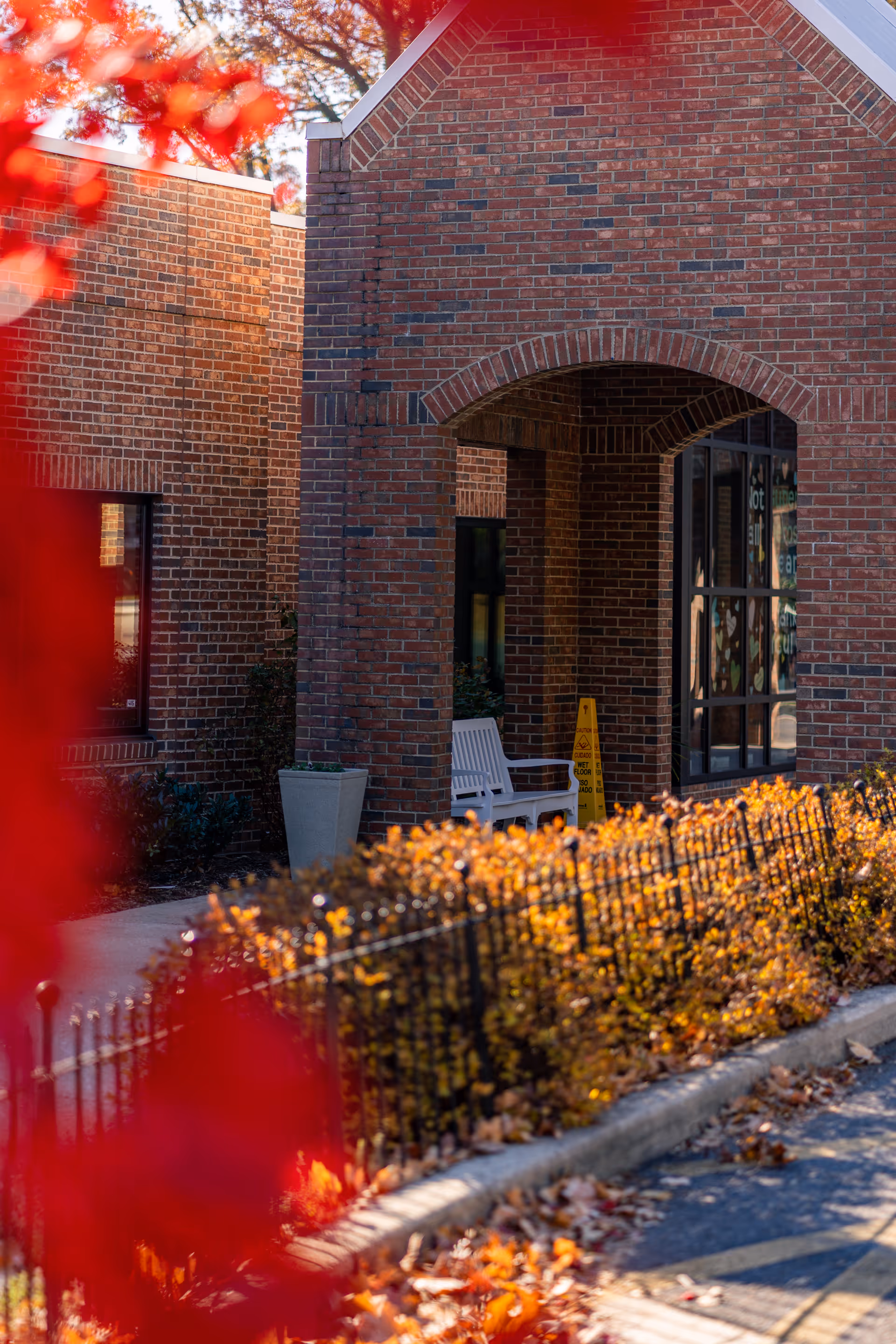 Exterior view of a brick building entrance with an arched doorway, a white bench, a yellow caution sign, and autumn foliage in the foreground.