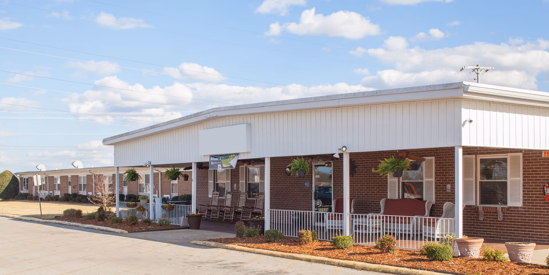 Exterior view of a single-story brick senior living facility with a covered porch featuring rocking chairs and hanging plants under a partly cloudy sky.