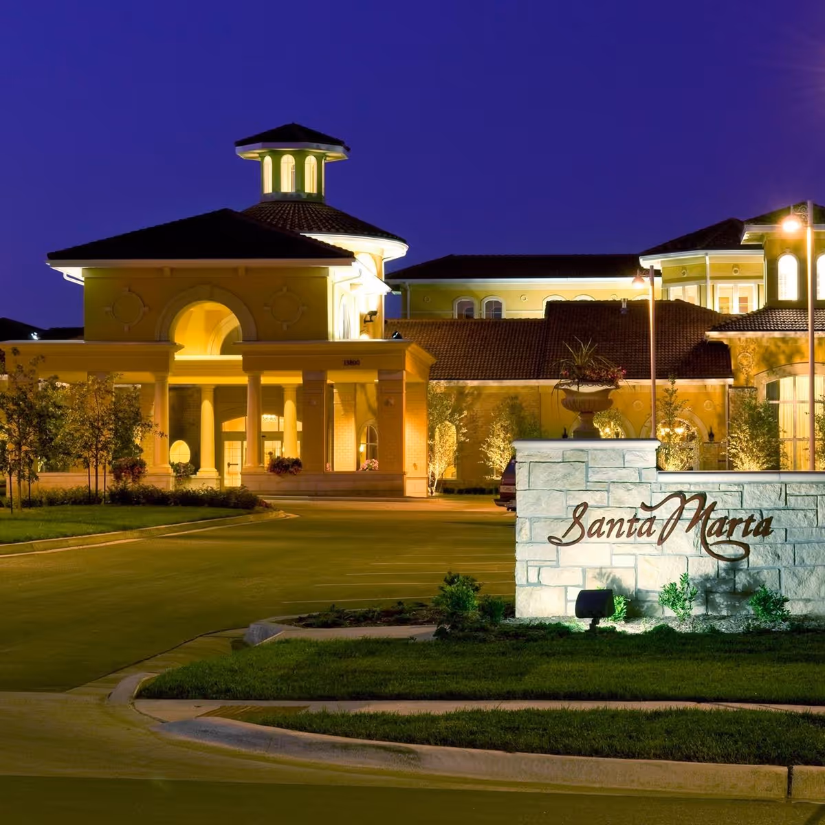 Night view of the illuminated entrance and exterior of the Santa Marta senior living facility with a stone sign displaying the facility name in front and well-maintained landscaping.