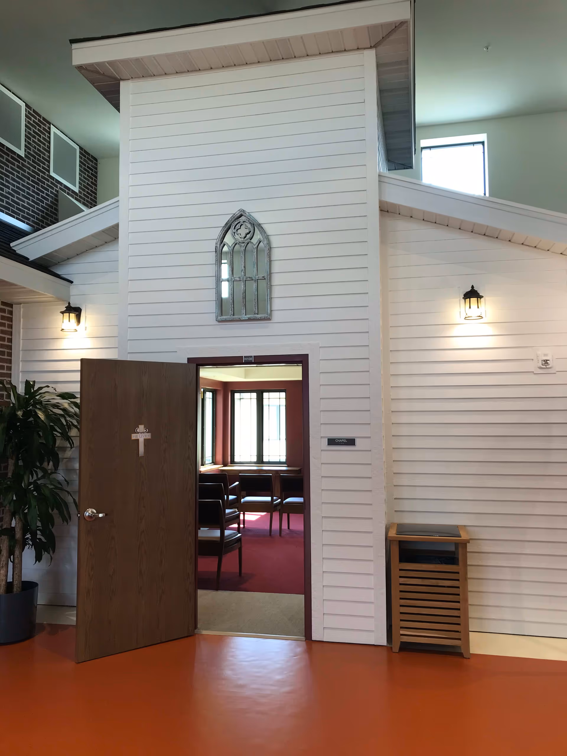 Interior view of a chapel entrance within a senior living facility. The chapel has white paneled walls with a decorative arched mirror above the door. The door is open, showing chairs arranged inside on a red carpet. There are two wall-mounted lantern-style lights on either side of the entrance and a potted plant to the left.