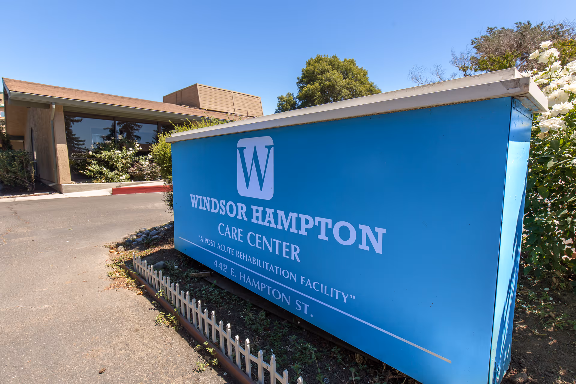 Exterior view of Windsor Hampton Care Center with a large blue sign displaying the facility's name and address, surrounded by greenery and a clear blue sky.