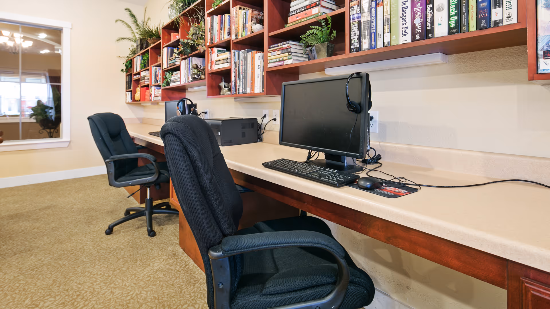 A row of computer workstations with office chairs beneath wall-mounted bookshelves filled with books and plants in a common area.