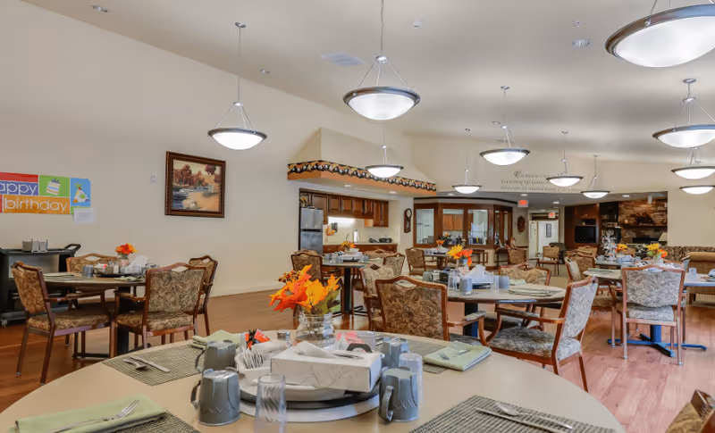 A spacious dining room in an assisted living facility with multiple round tables set with placemats, napkins, cups, and utensils. The room features wooden chairs with patterned upholstery, warm lighting from ceiling fixtures, and a small kitchen area in the background. There are autumn-themed floral centerpieces on the tables and a 'Happy Birthday' sign on the wall.
