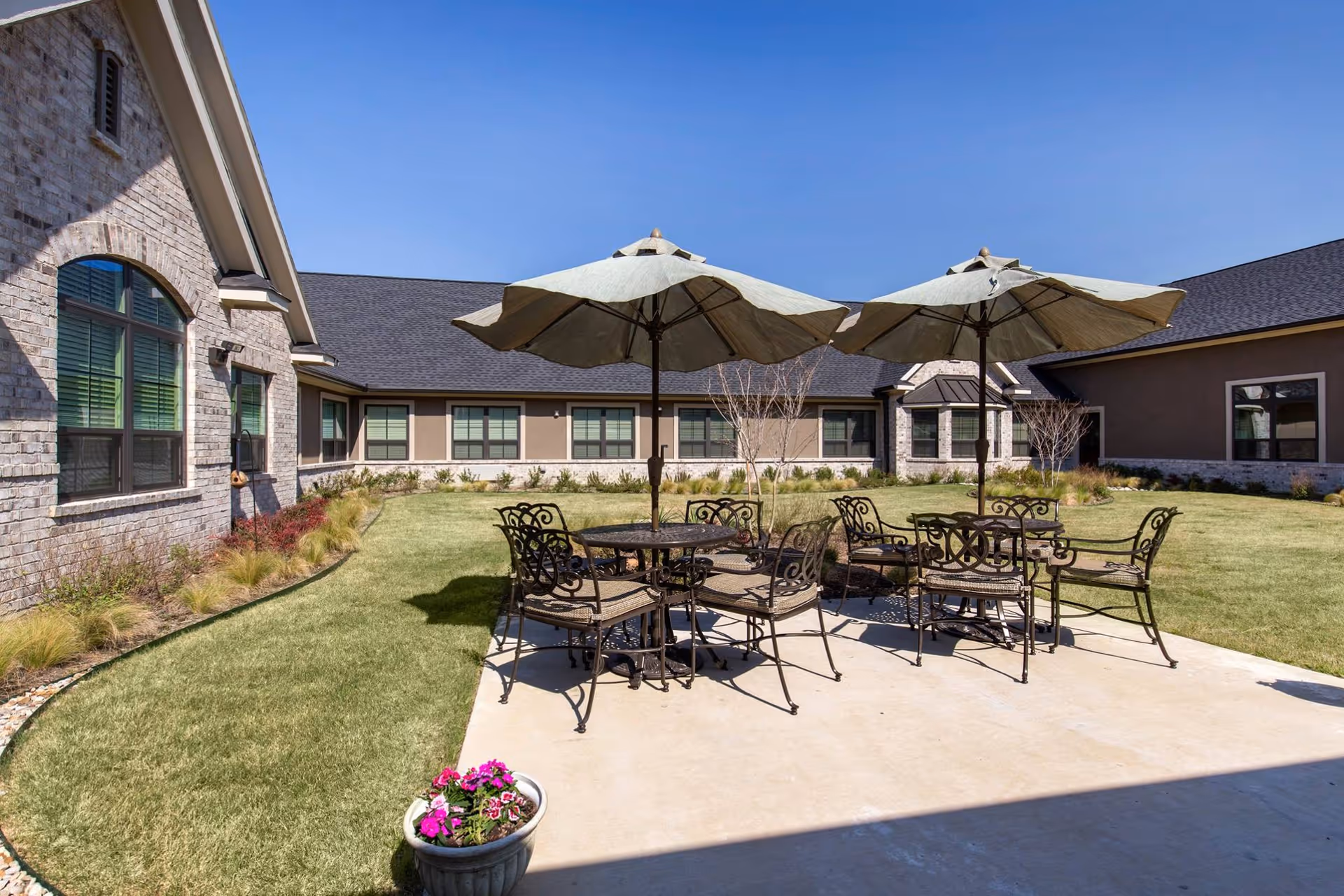Outdoor patio area at Briarview Senior Living with metal tables and chairs under large beige umbrellas on a concrete surface, surrounded by a grassy courtyard and buildings with windows under a clear blue sky.