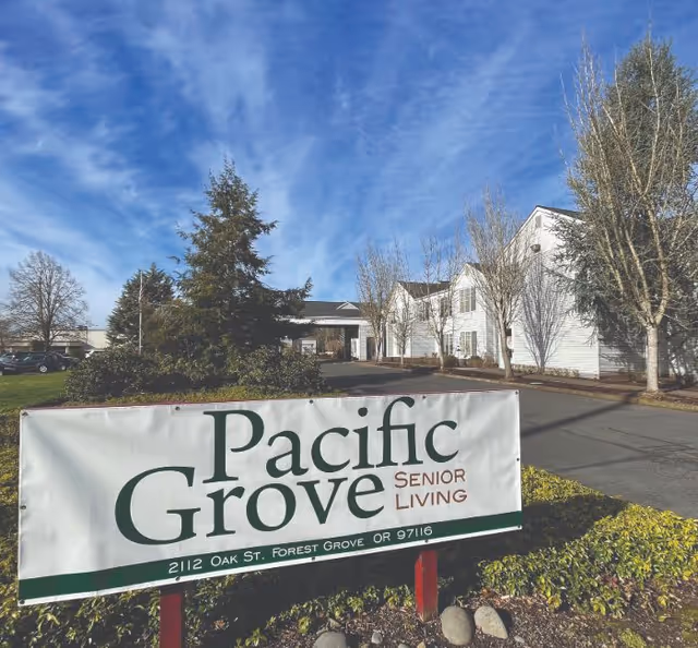 Exterior view of Pacific Grove Senior Living facility with a large sign in the foreground displaying the facility's name and address. The building is white with multiple windows, surrounded by leafless trees and greenery under a partly cloudy blue sky.
