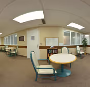 Interior community room with round tables and chairs, a small bookshelf, bulletin board, windows and fluorescent ceiling lights.