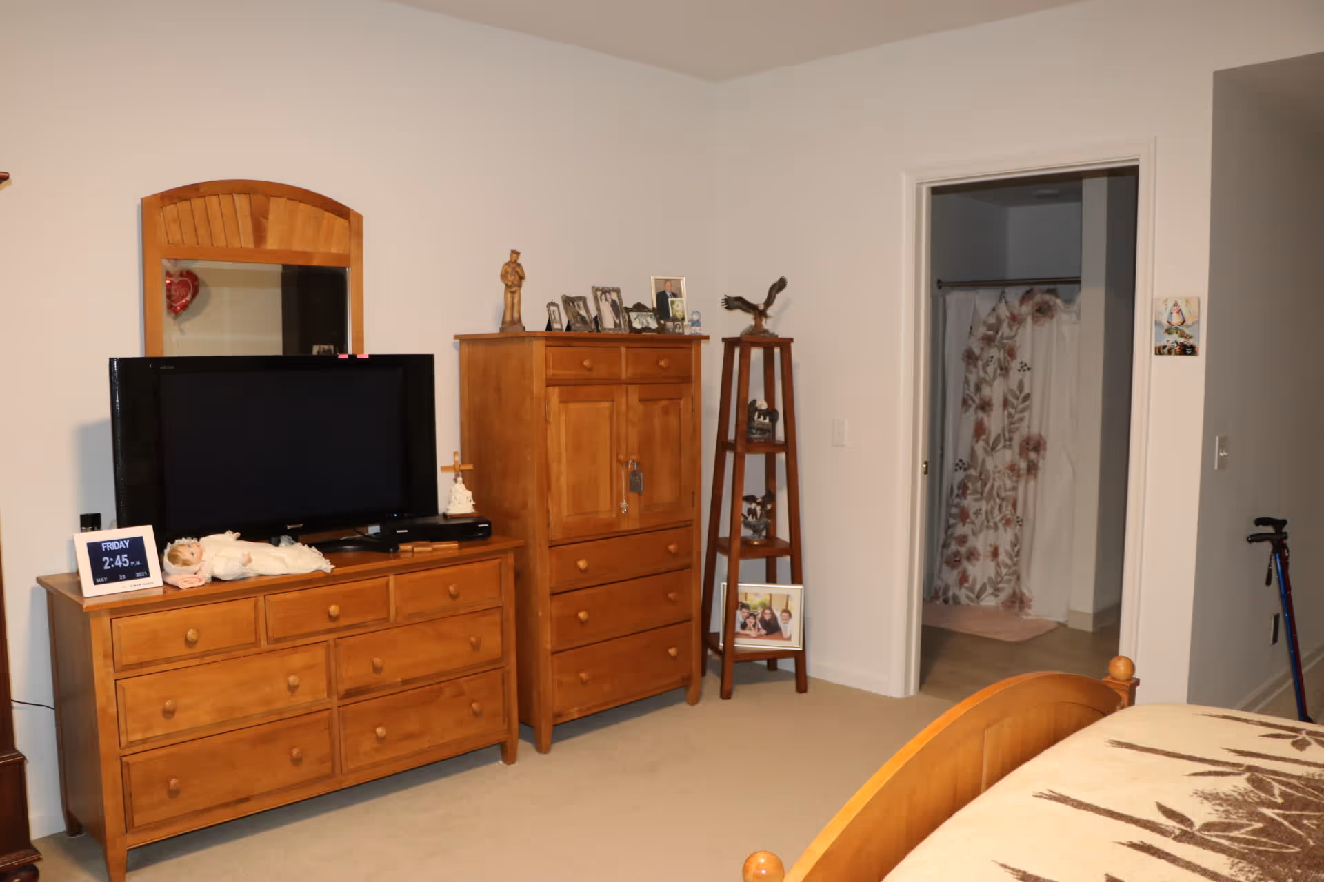 A bedroom with wooden furniture including a dresser with a TV on top, a tall chest of drawers, and a corner shelf with decorative items and framed photos. There is a bed partially visible in the foreground with a patterned blanket. An open door reveals a bathroom with a floral shower curtain. A walking cane is leaning against the wall near the bathroom door.