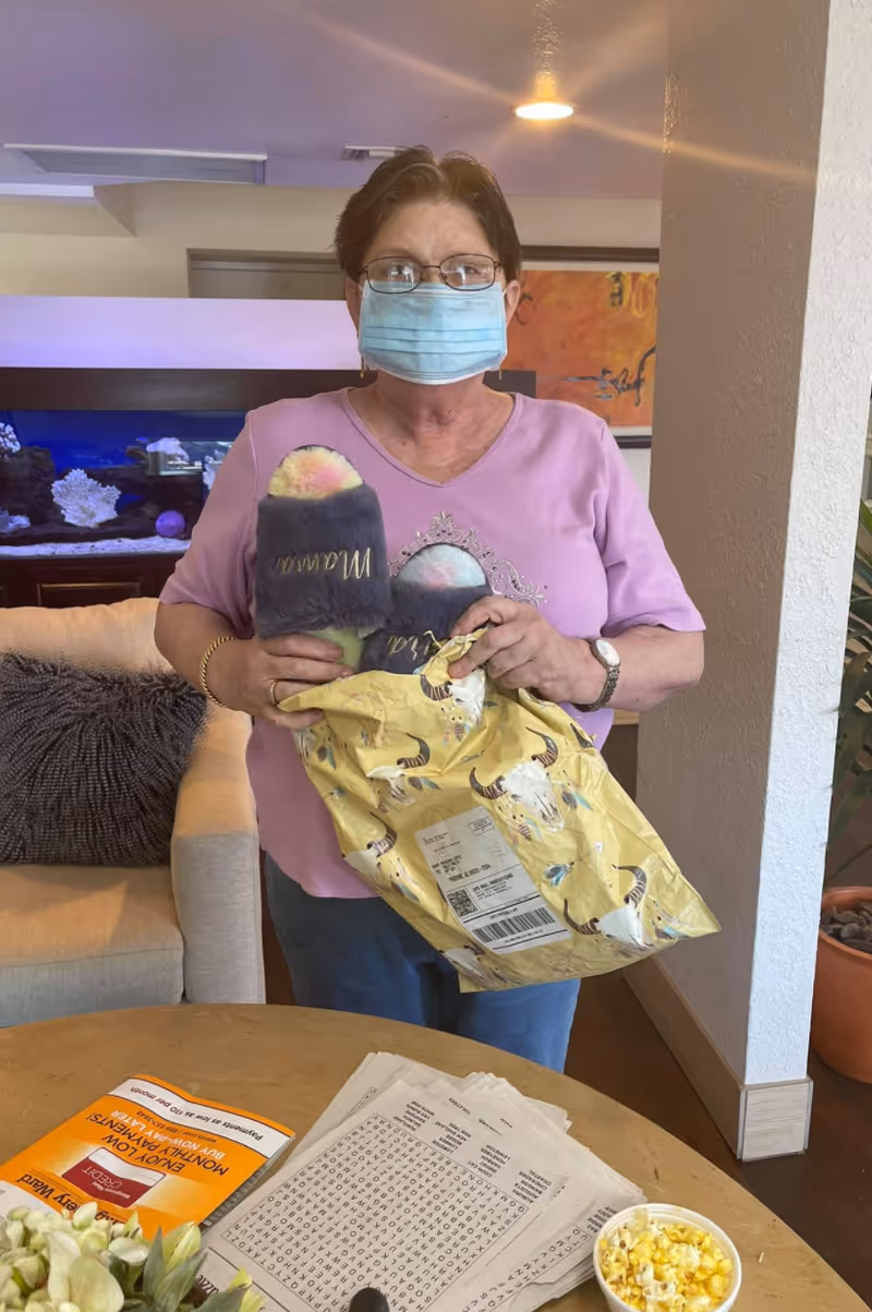 An elderly woman wearing glasses and a face mask stands indoors holding a pair of fuzzy slippers and a yellow package. She is wearing a pink shirt and standing near a table with papers, a word search puzzle, and a small bowl of popcorn. Behind her is a beige couch with a dark pillow and an aquarium.