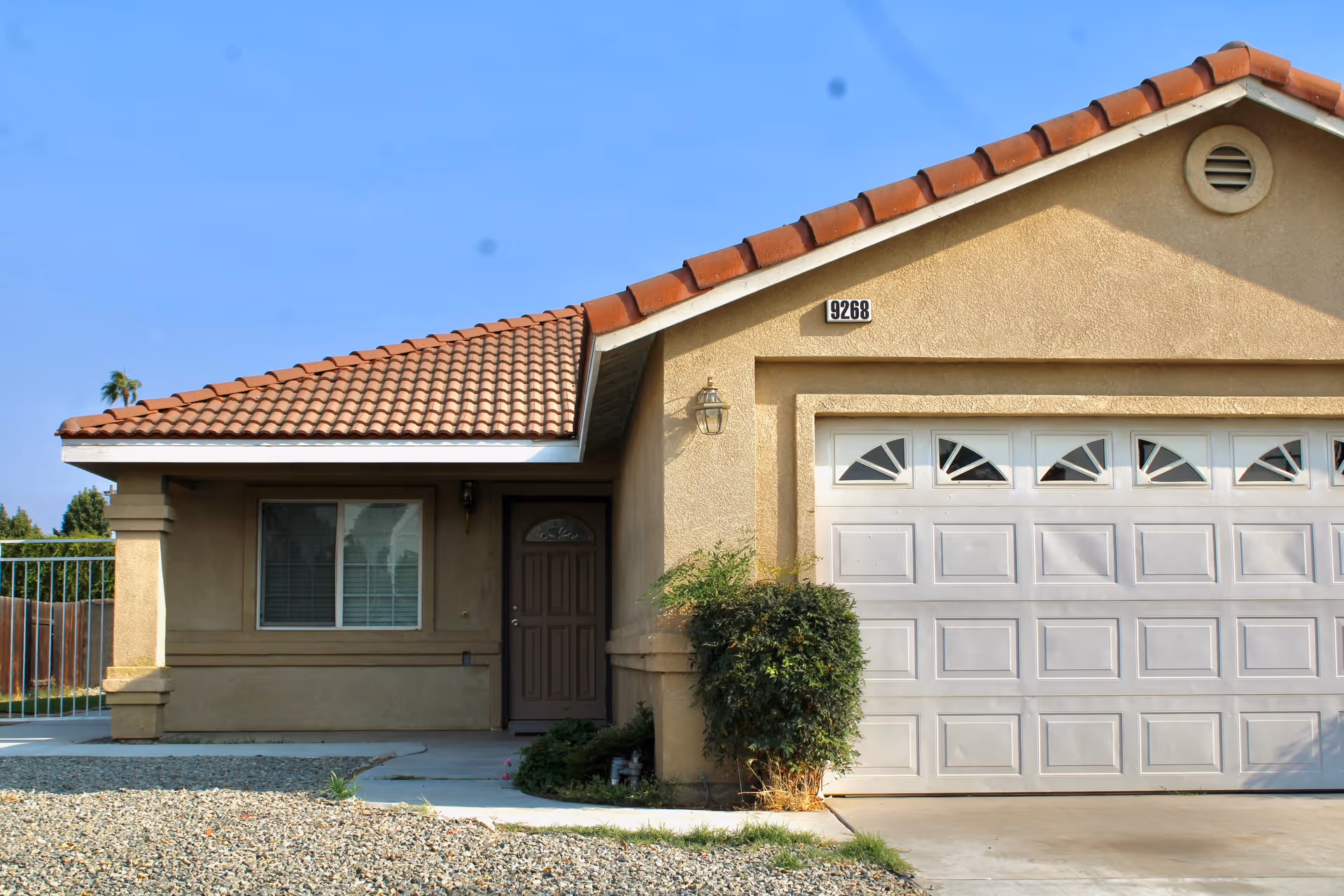 Exterior view of a single-story house with a beige stucco finish and a red tile roof. The house features a white garage door with decorative windows, a brown front door, a window with closed blinds, and a small bush near the entrance. The house number 9268 is displayed above the garage door. The sky is clear and blue.