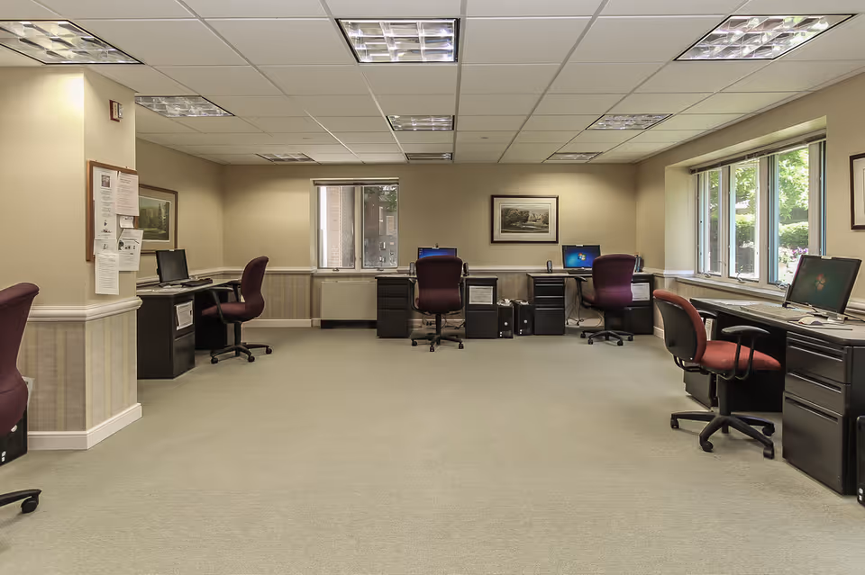 A spacious office area with multiple computer workstations along the walls. Each workstation has a desk, a computer, and an office chair with maroon upholstery. The room has beige walls, a light-colored carpet, and a drop ceiling with fluorescent lighting. There are windows on two walls letting in natural light, and framed artwork is hung on the walls.