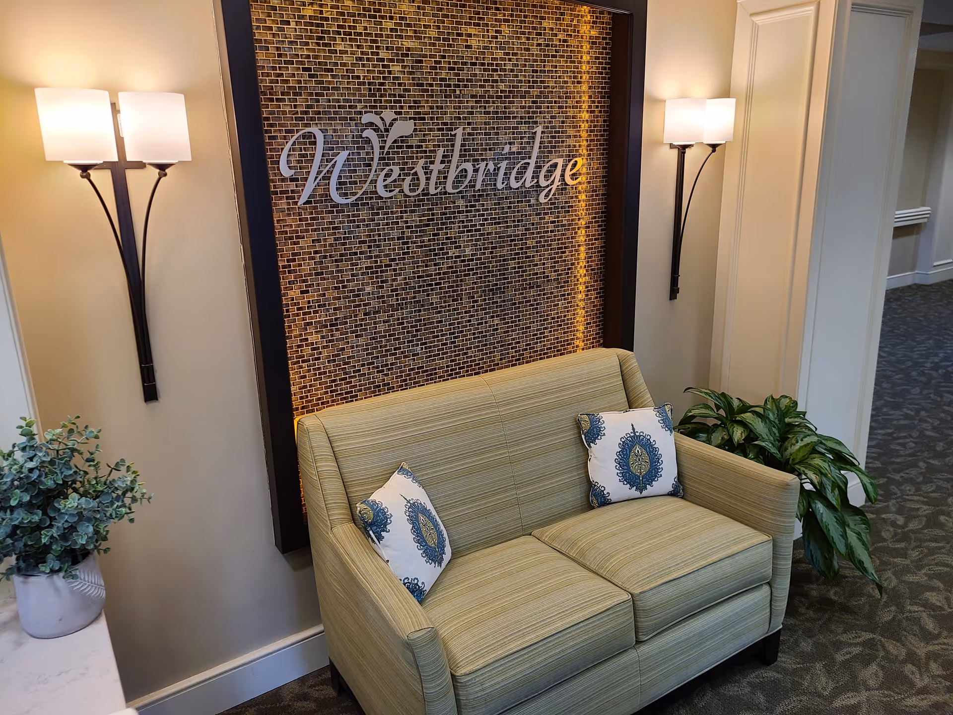 A cozy seating area in a hallway with a light green striped loveseat adorned with two decorative pillows featuring blue and green patterns. Behind the loveseat is a wall with a mosaic tile design and the word 'Westbridge' displayed prominently. Two wall-mounted lamps with white shades provide warm lighting. There are also two potted plants on either side of the loveseat.