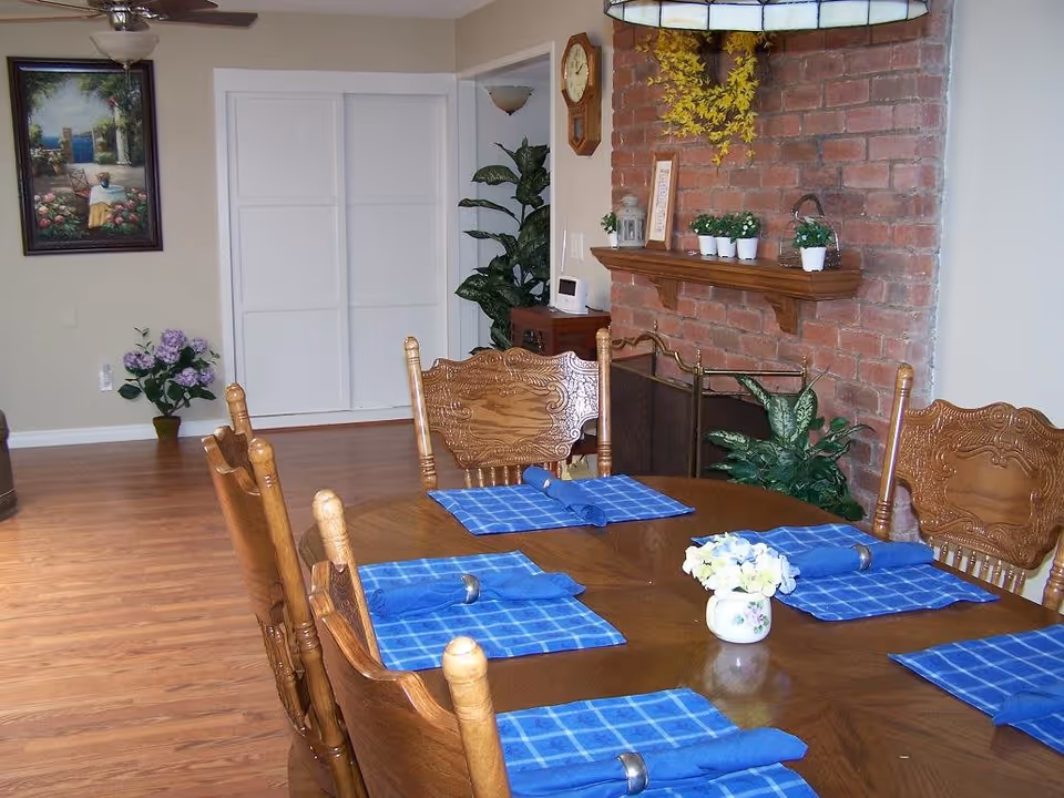 A dining room with a wooden table set for six people with blue checkered placemats and blue napkins with silver napkin rings. The room features wooden chairs with carved backs, a brick fireplace with a wooden mantel decorated with small potted plants and a framed picture. There is a wall clock above the fireplace and a large green plant next to it. The floor is wooden, and there is a framed painting and a potted plant against the beige wall in the background.