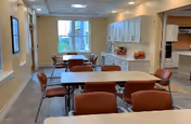 A bright dining room with several tables and orange cushioned chairs arranged neatly. There is a large window at the far end letting in natural light, and white cabinets with some decorative items on top along the right wall. The room has a clean, welcoming atmosphere.