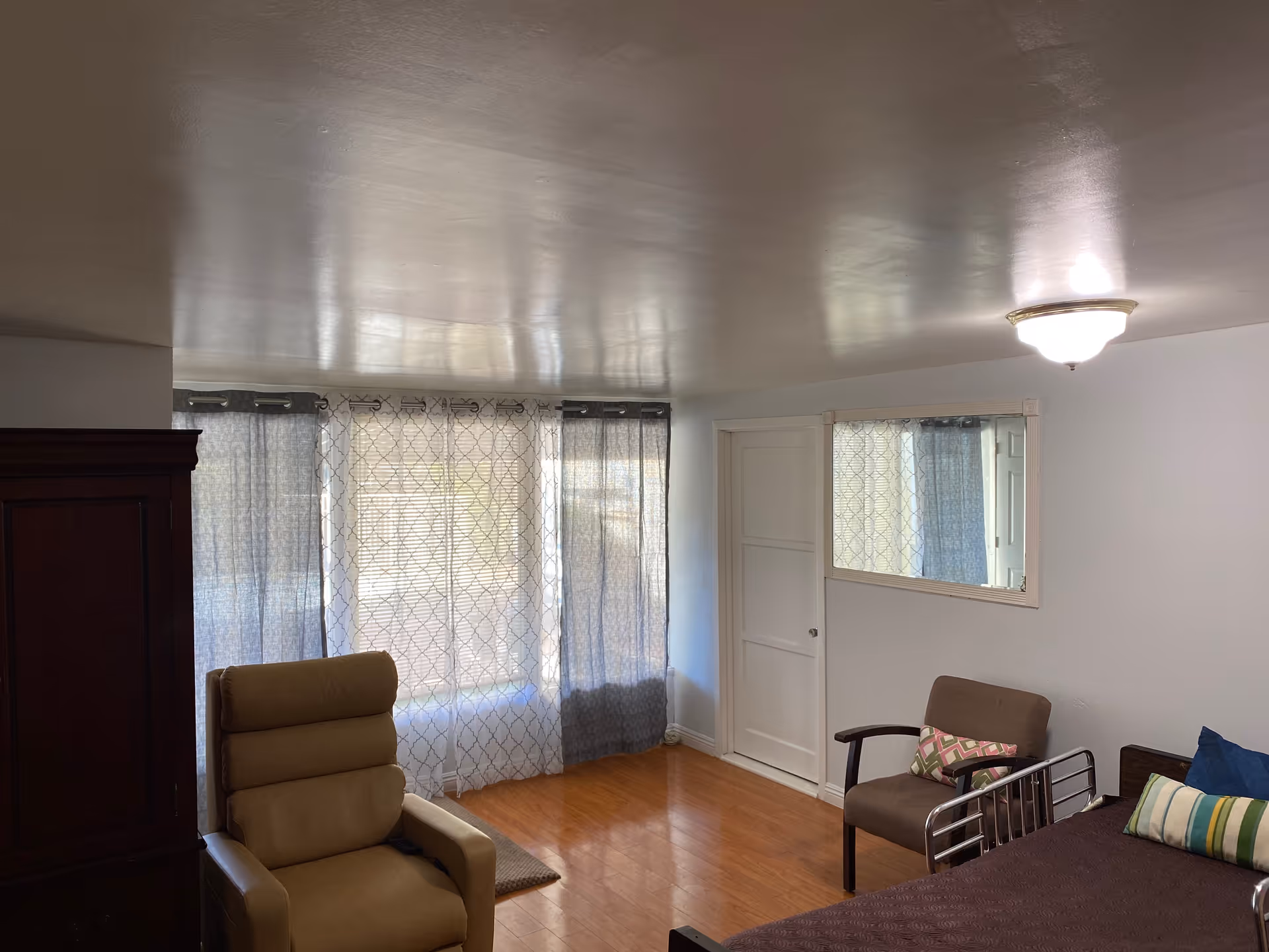 A small room with wooden flooring featuring a brown recliner chair, a brown armchair with a patterned cushion, a bed with pillows, a large window covered with sheer and dark curtains, a wooden cabinet, a wall mirror, and a ceiling light fixture.