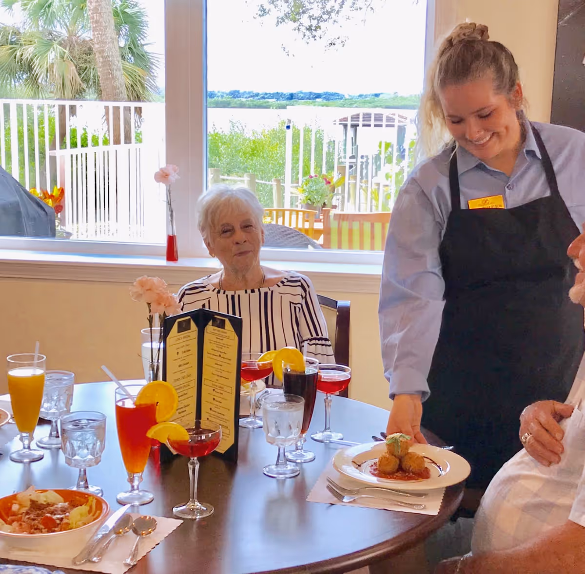 An elderly woman sitting at a dining table with various drinks and a menu in front of her, while a smiling waitress serves a plate of food to another person at the table. The background shows a large window with a view of outdoor greenery and a patio area.