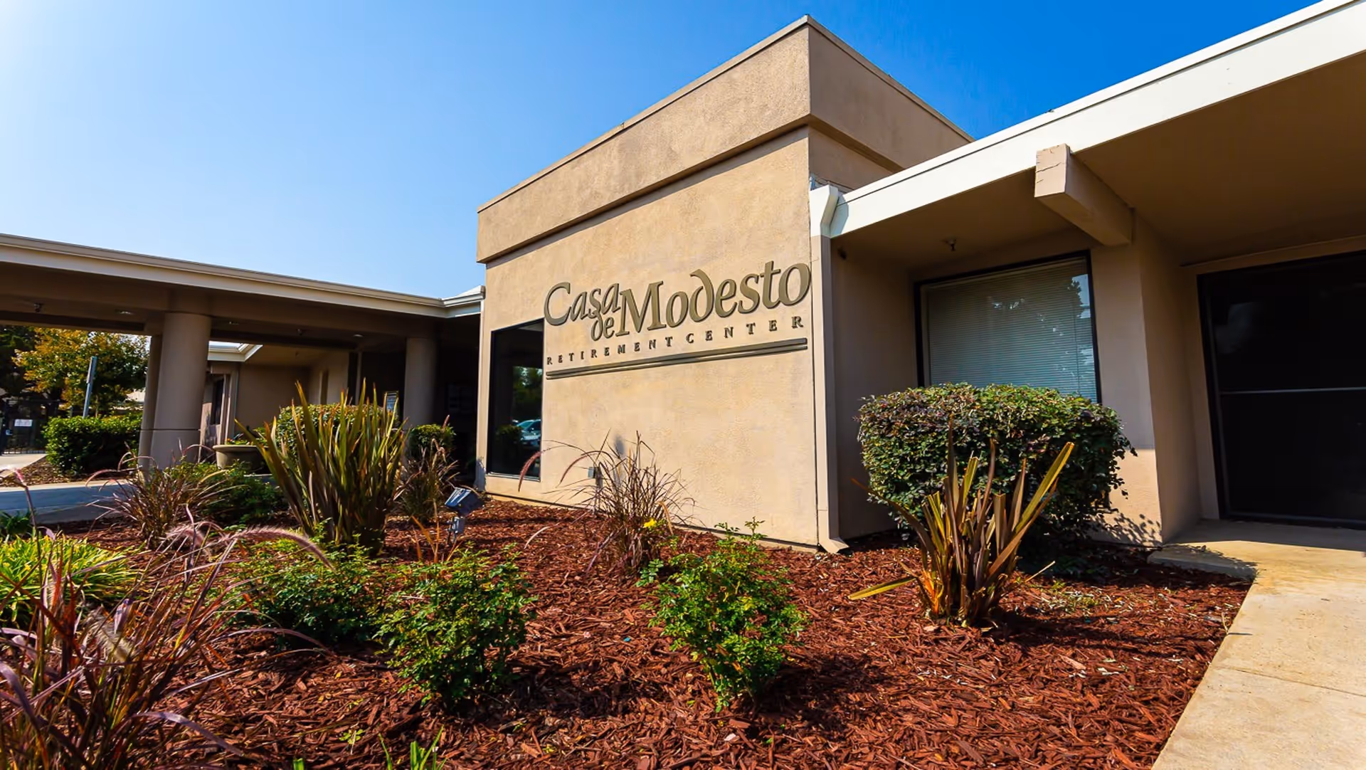 Exterior view of Casa de Modesto Retirement Center building with beige walls, a window, and landscaped garden with various plants and mulch in front under a clear blue sky.