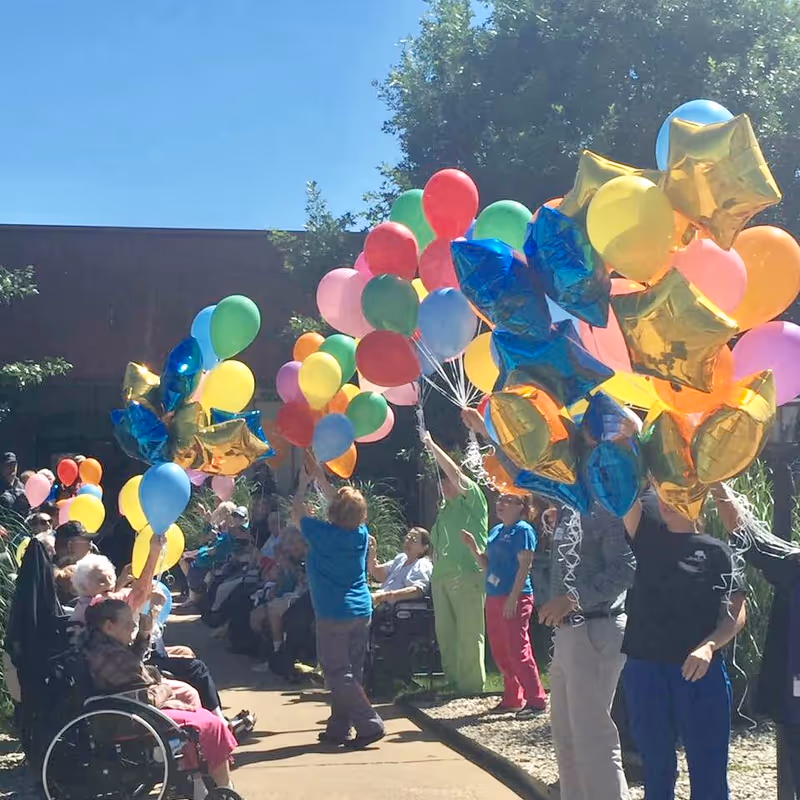 A group of elderly people and staff members outdoors holding colorful balloons, including star-shaped gold and blue balloons, on a sunny day at a nursing and rehabilitation facility.