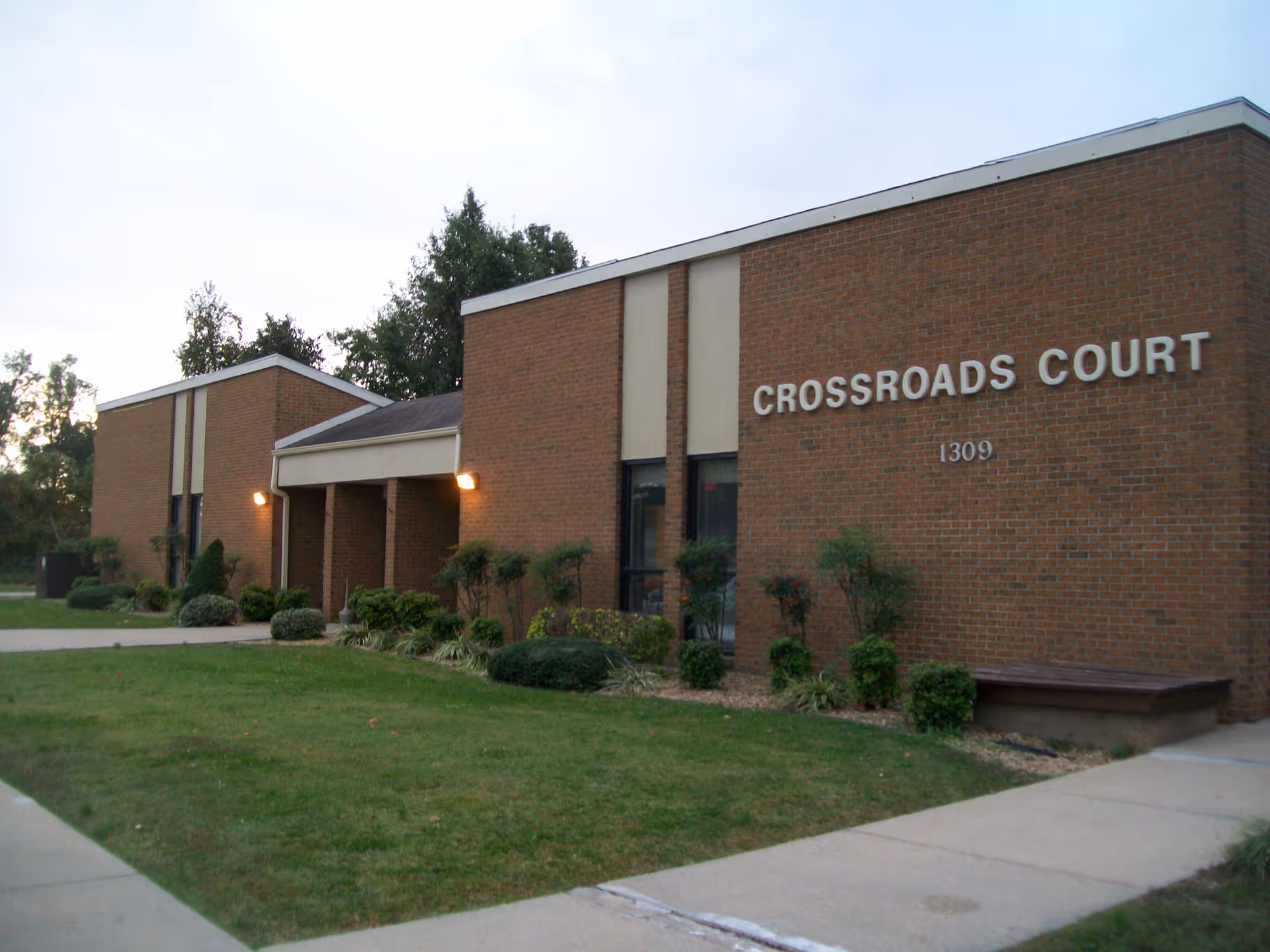 Exterior view of a brick building with the name 'Crossroads Court' and the number '1309' displayed on the front. The building has a small covered entrance with lights on either side, surrounded by neatly trimmed bushes and a green lawn.