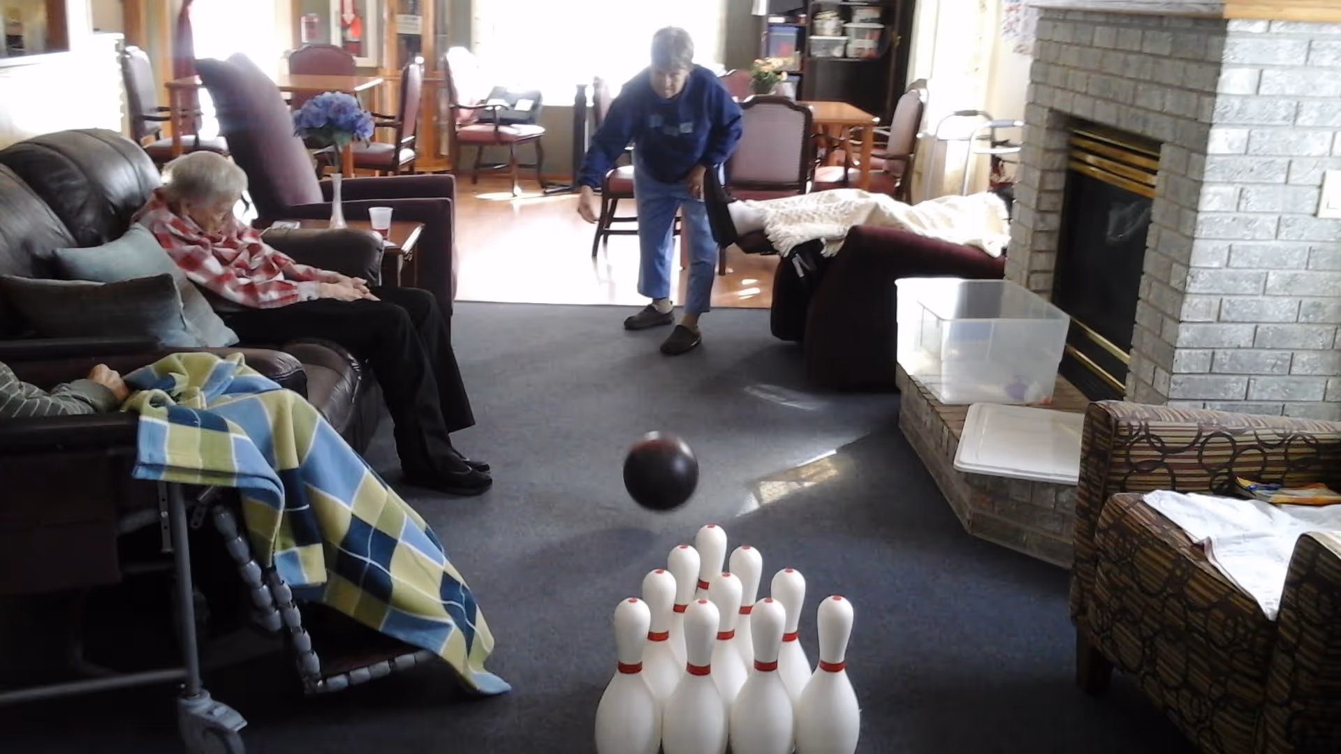 An elderly woman is bowling with a black ball towards white bowling pins set up on a carpeted floor in a living room area. Two other elderly individuals are seated nearby, one covered with a blue and green checkered blanket. The room has comfortable chairs, a fireplace, and a dining area in the background.