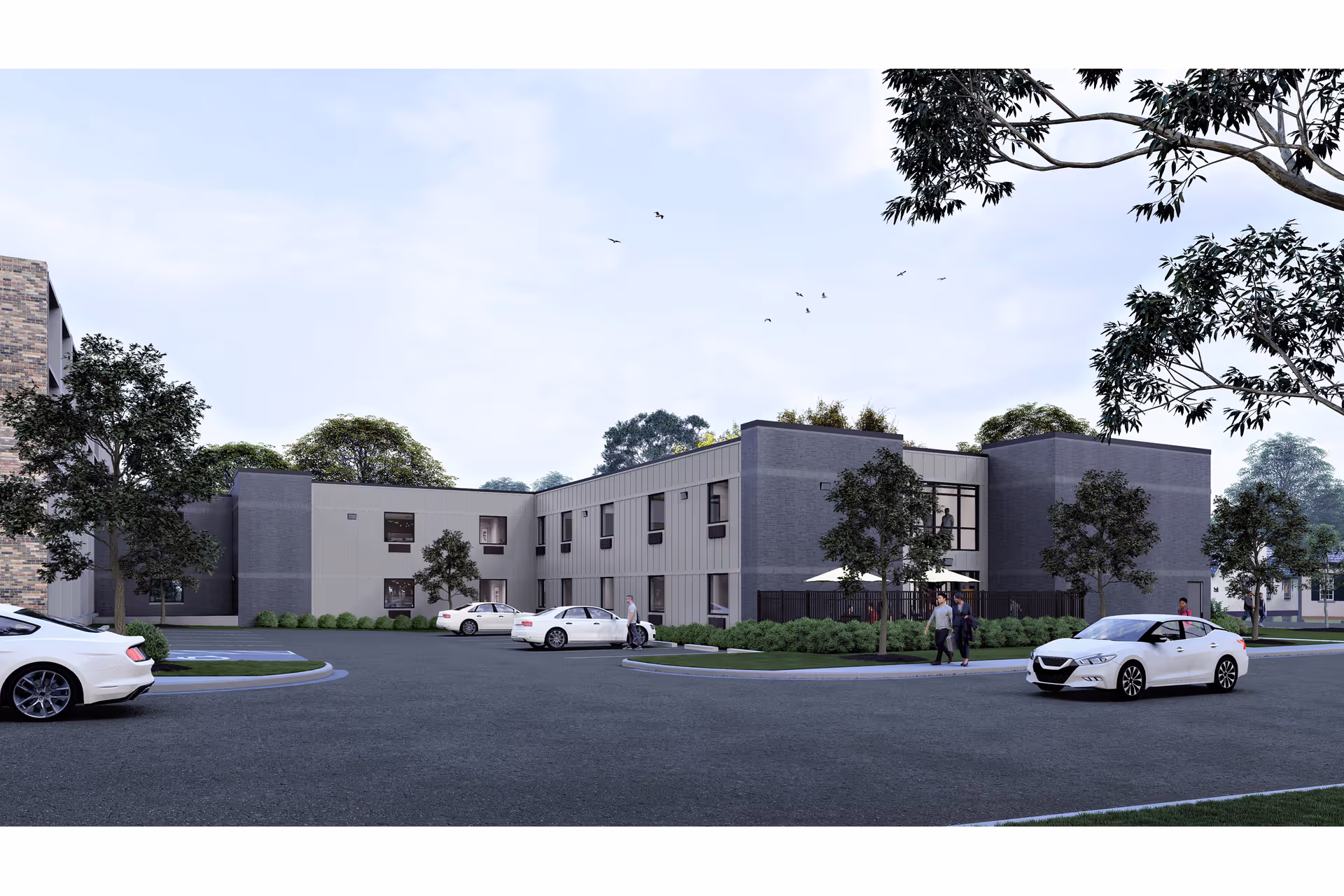 Exterior view of a modern two-story supportive living and memory care facility with several white cars parked in front, a few people walking near the entrance, and trees surrounding the building under a partly cloudy sky.