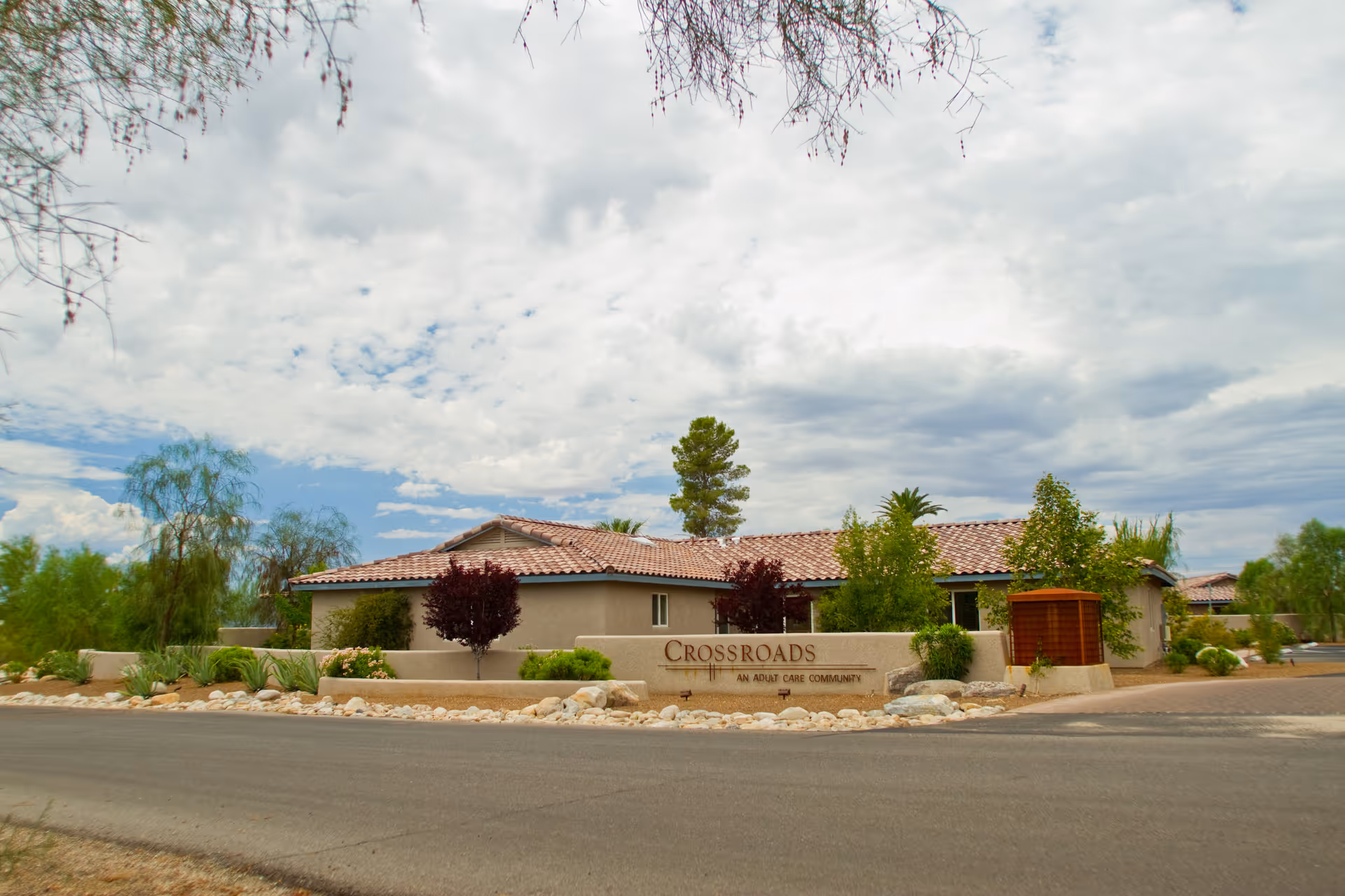 Exterior view of Crossroads Assisted Living, a single-story building with a tiled roof surrounded by trees and landscaping under a partly cloudy sky. A stone sign in front reads 'Crossroads An Adult Care Community.'