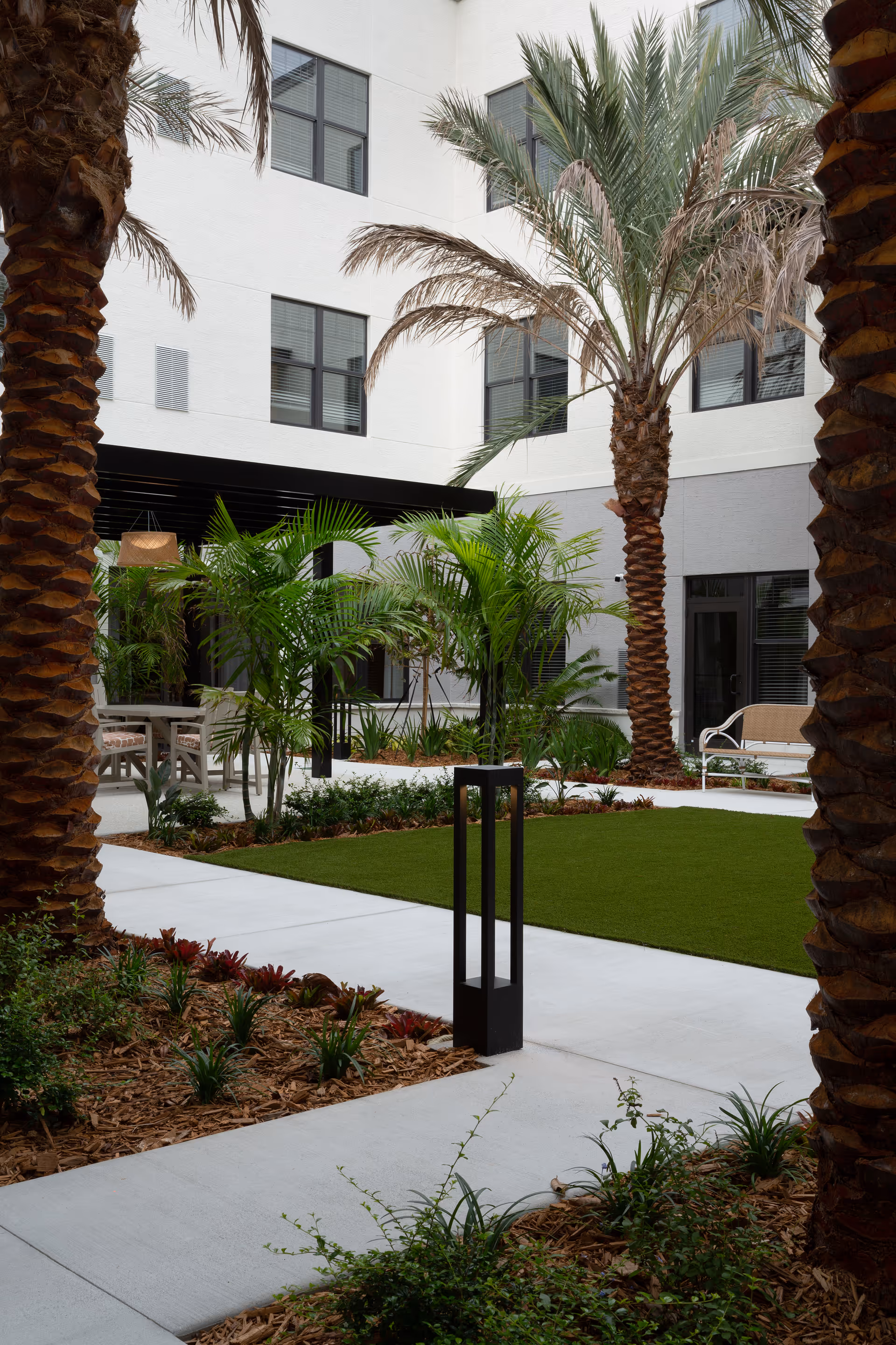 Outdoor courtyard area at The Palms at Plantation featuring palm trees, green plants, a manicured lawn, concrete walkways, and seating areas with a white building in the background.