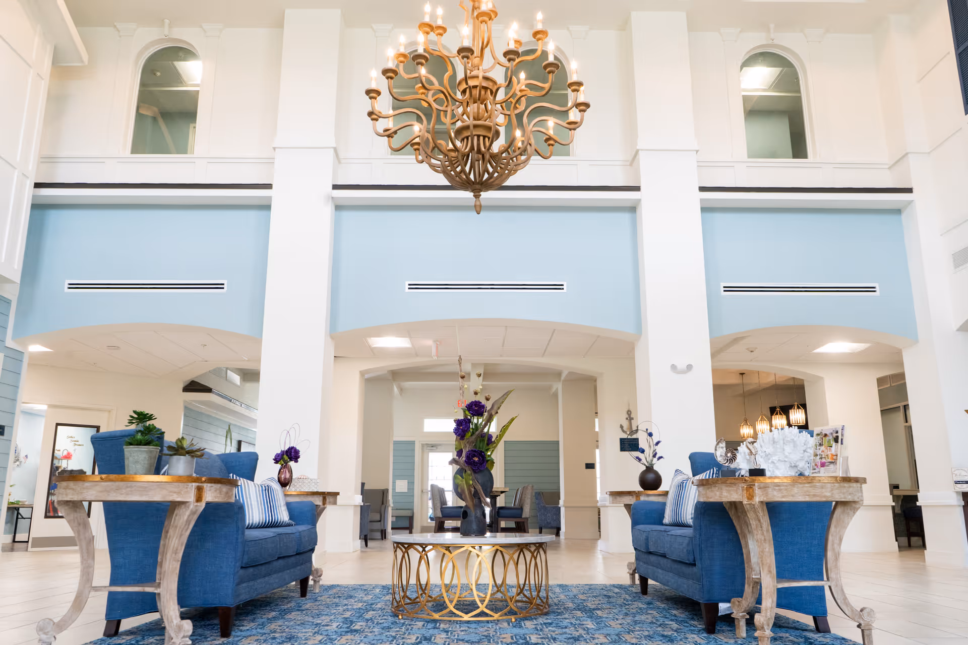 A spacious and elegant living room area with high ceilings and a large ornate chandelier hanging from the ceiling. The room features two blue upholstered sofas facing each other with wooden side tables on each side, decorated with small plants and vases. A round coffee table with a gold geometric base and a floral centerpiece is placed between the sofas on a blue patterned rug. The walls are painted white with light blue accents, and there are large archways and windows letting in natural light.