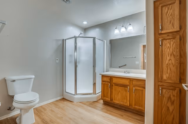 A clean bathroom featuring a white toilet, a corner glass shower with frosted doors, a wooden vanity with a white countertop and sink, a large mirror above the vanity with three light fixtures, and wooden cabinetry on the right side. The floor has a light wood finish and the walls are painted light gray.