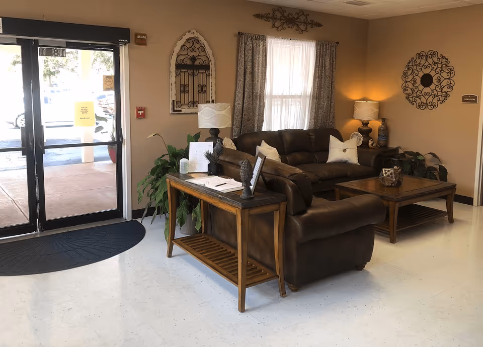 A cozy seating area inside a facility with brown leather sofas, a wooden coffee table, and a side table with a lamp and decorative items. There is a large window with curtains, wall decorations, and a glass door leading outside with a black mat on the floor.