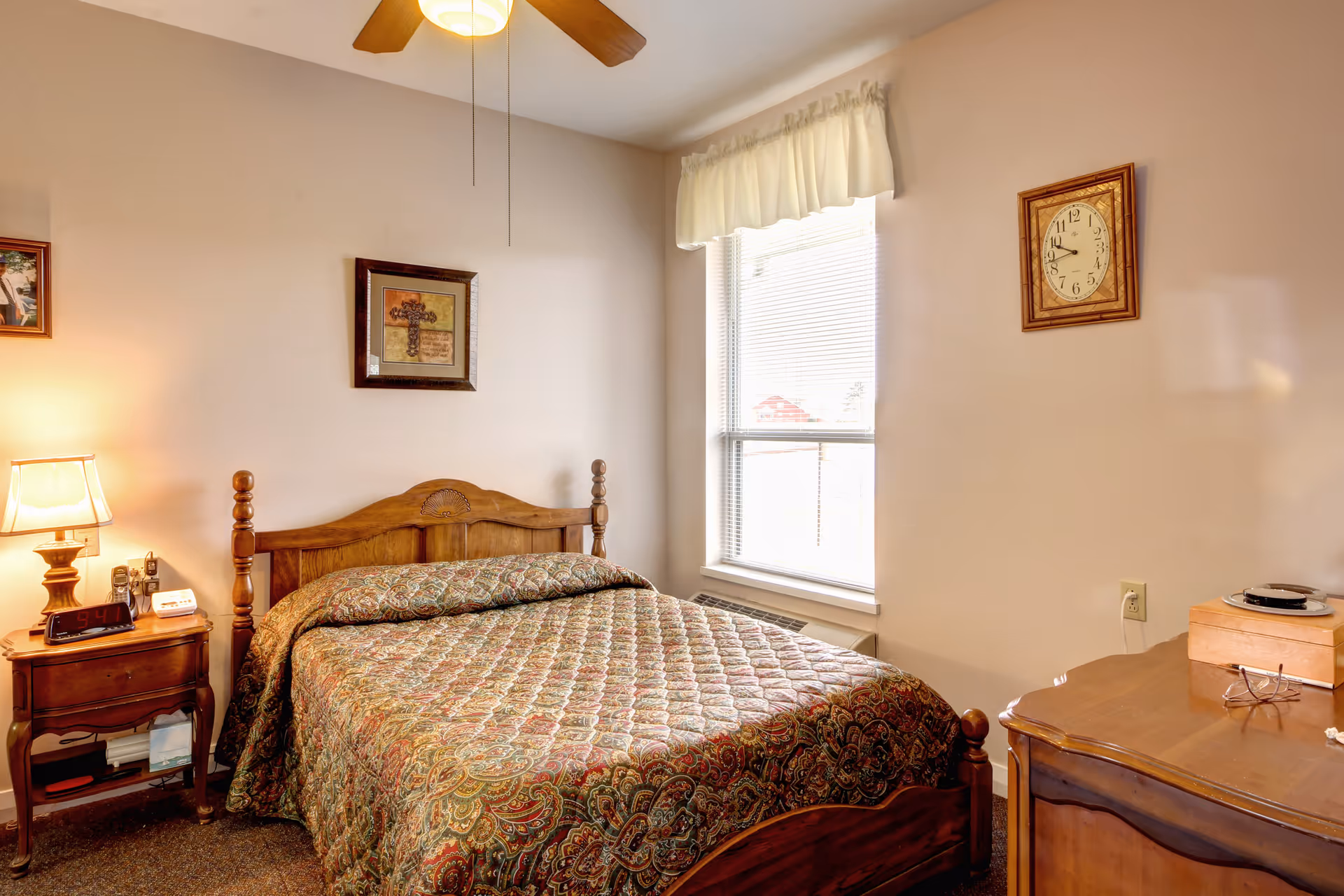 A cozy bedroom with a wooden bed frame and a patterned quilt. There is a wooden nightstand with a lamp, clock, and phone on the left side of the bed. A window with blinds and a white valance lets in natural light. On the right side, there is a wooden dresser with a pair of glasses and a wooden box on top. The walls are decorated with a framed picture and a clock.